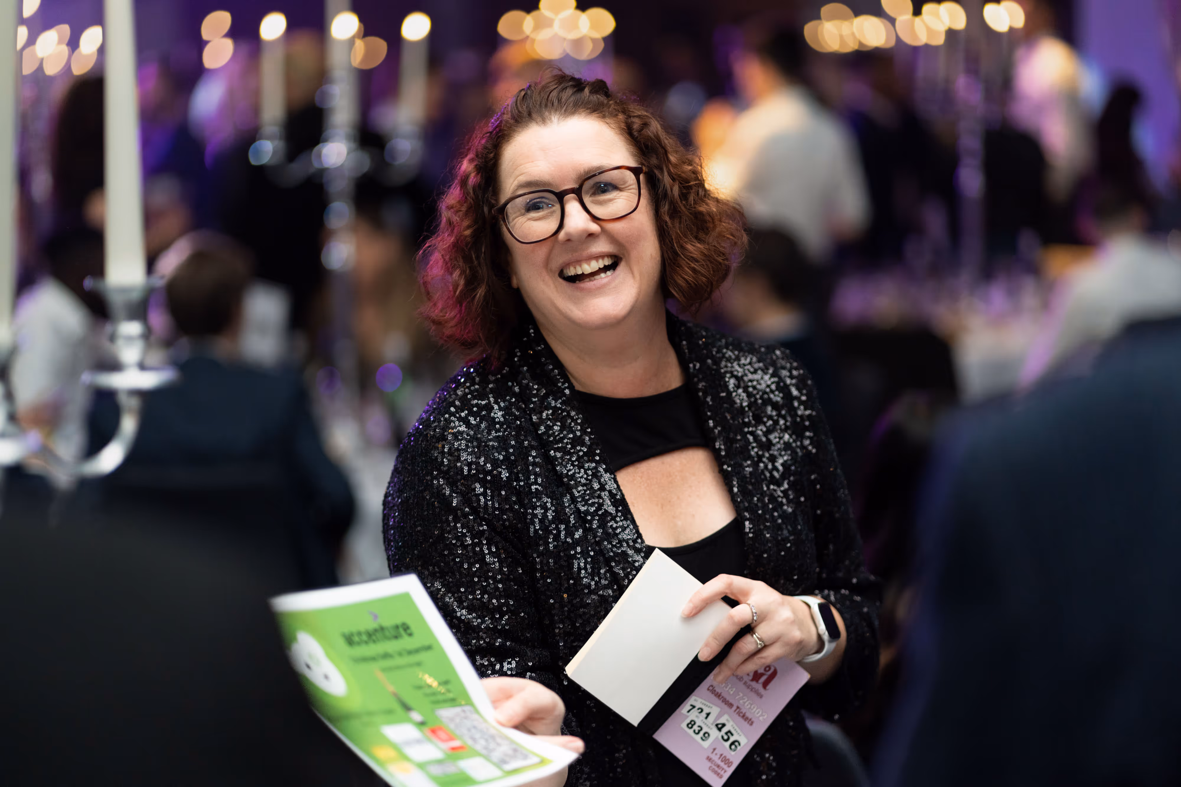 Smiling woman with glasses holds papers at a formal event with dim lights and people in the background.