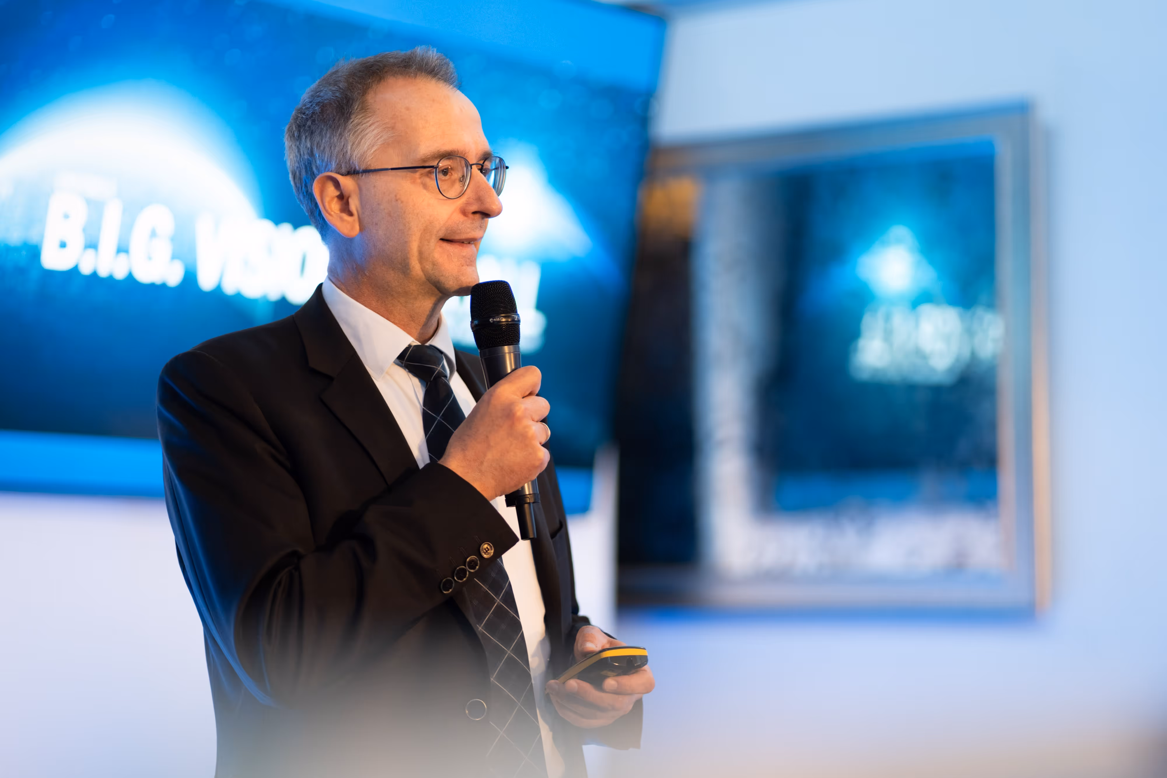 Man in a suit and glasses speaking into a microphone at an event with a screen displaying "B.I.G. VISION" behind him.