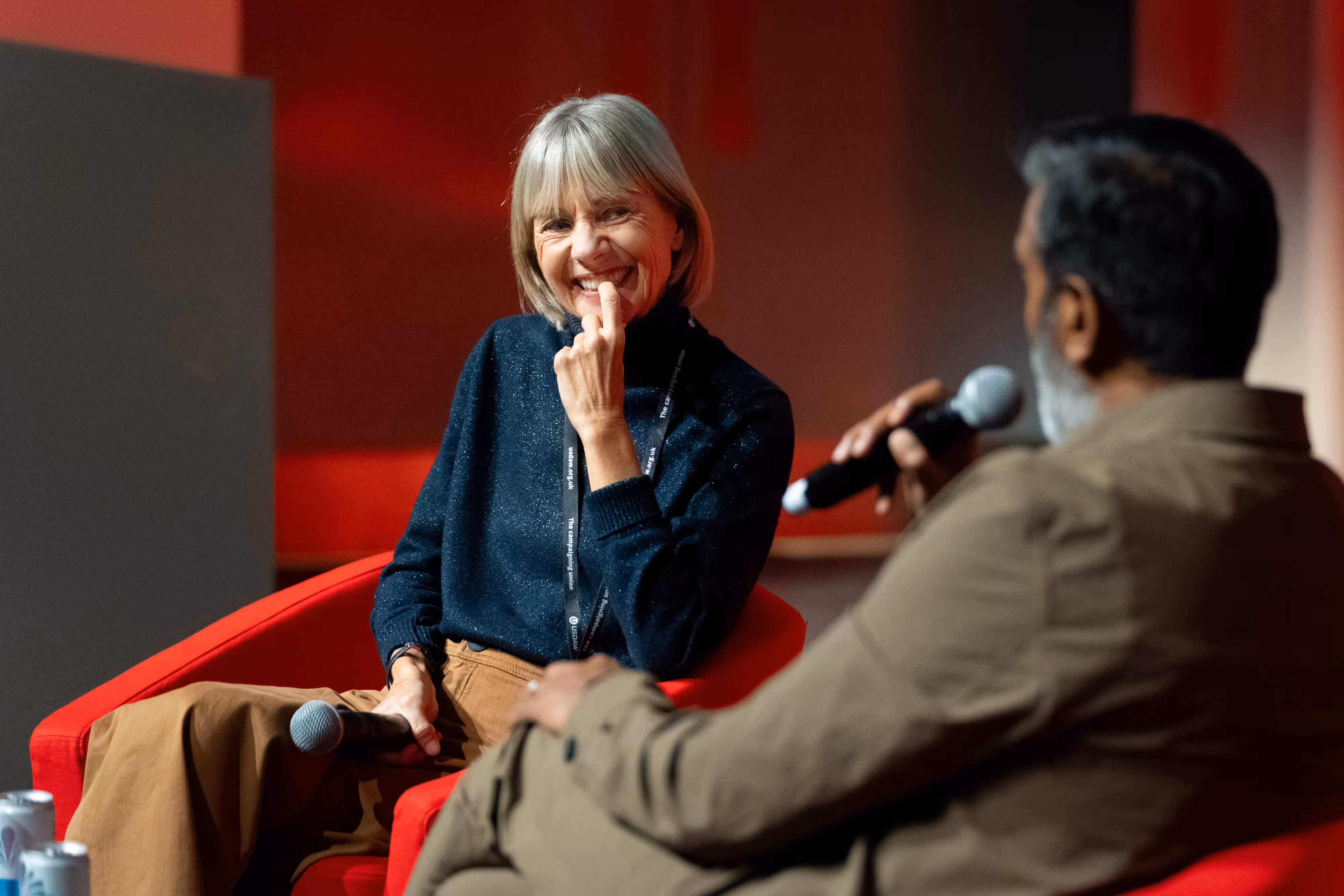 A smiling woman listens to a man speaking into a microphone during an indoor event with red lighting.