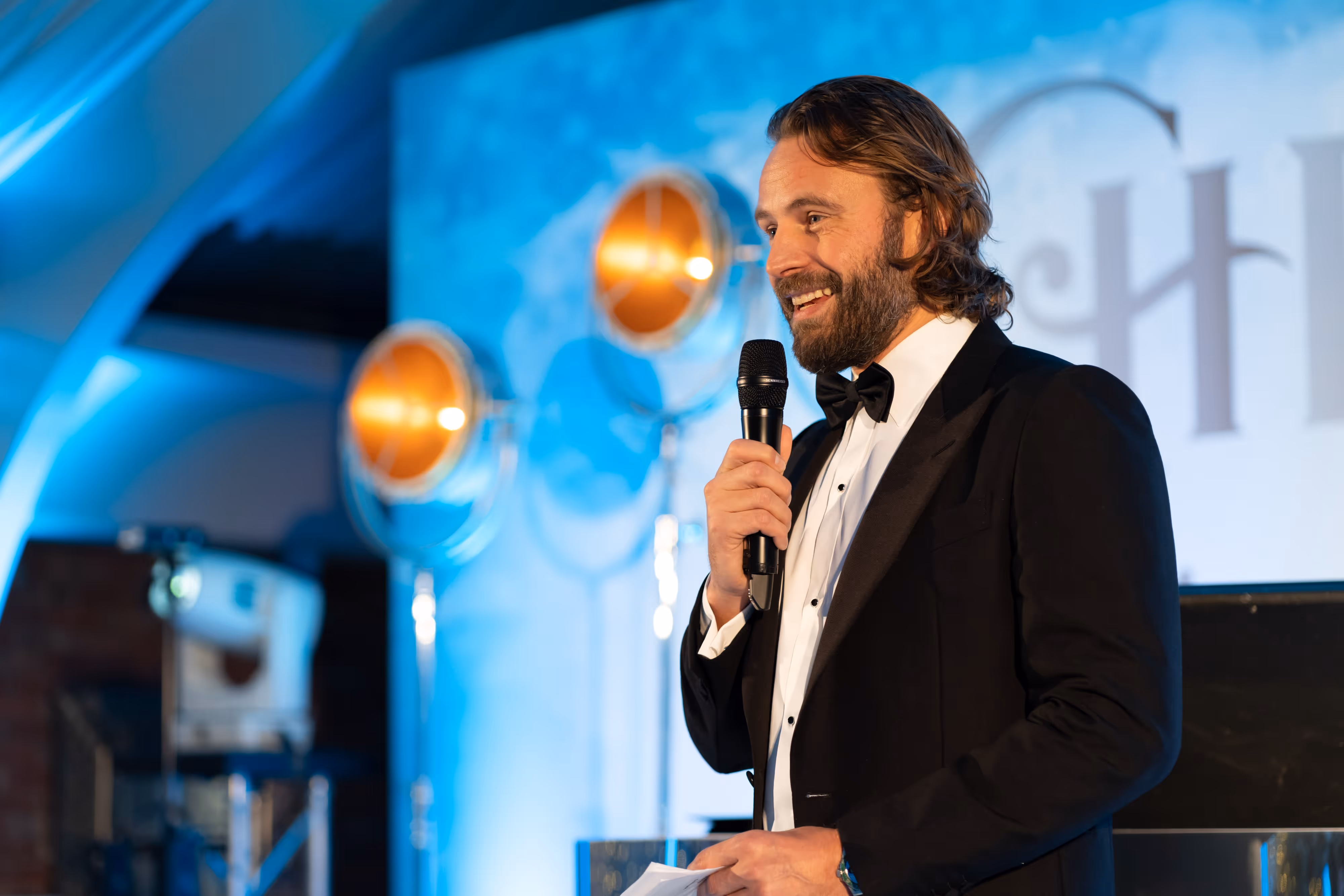 Man in a dinner jacket holding a microphone and speaking at an event with blue lighting in the background.