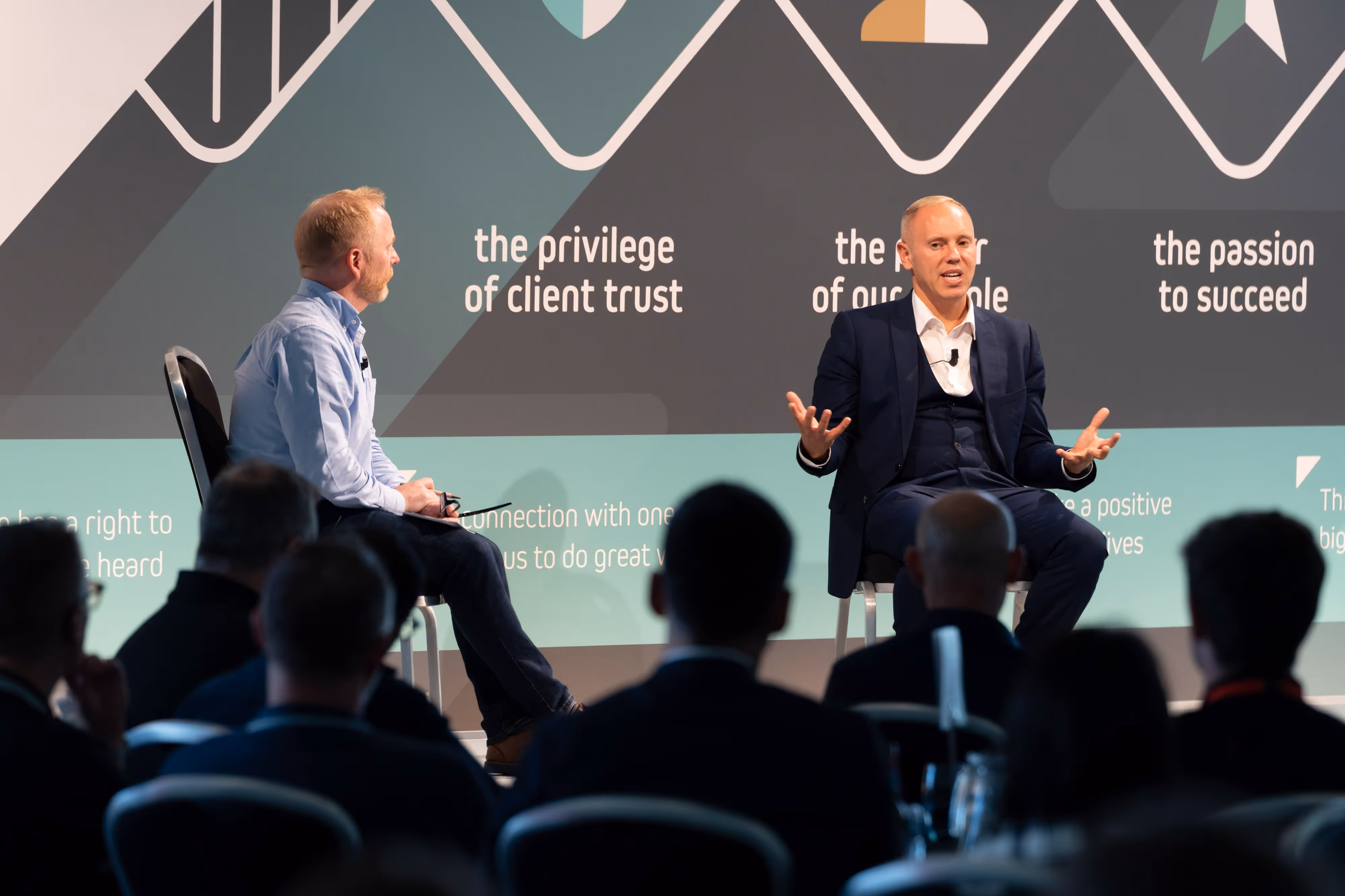 Two men sit on stage in conversation at a business event, facing an audience with a graphic backdrop behind them.