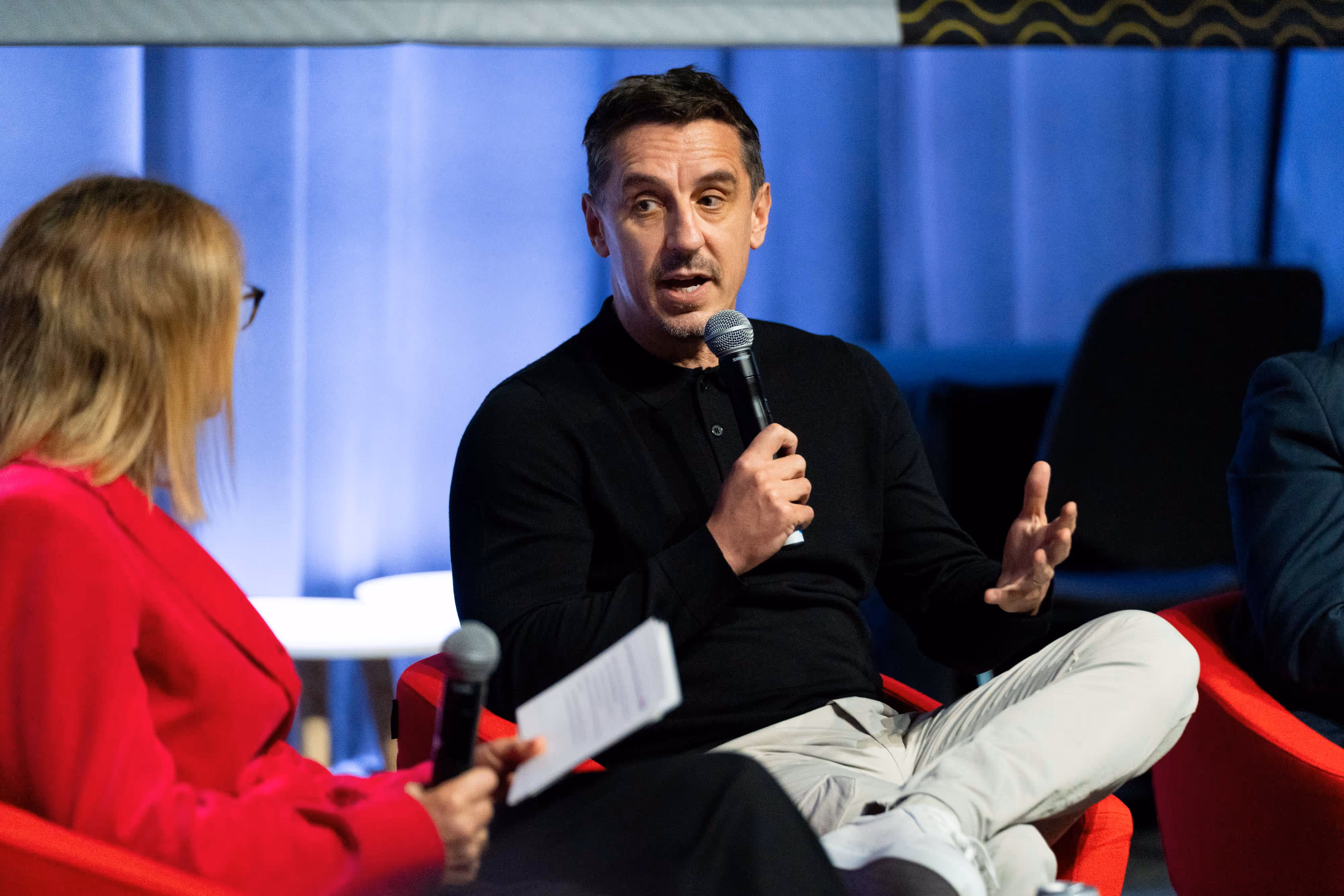 Gary Neville speaking into microphone while sitting on a red chair, woman in red jacket listens and holds papers.