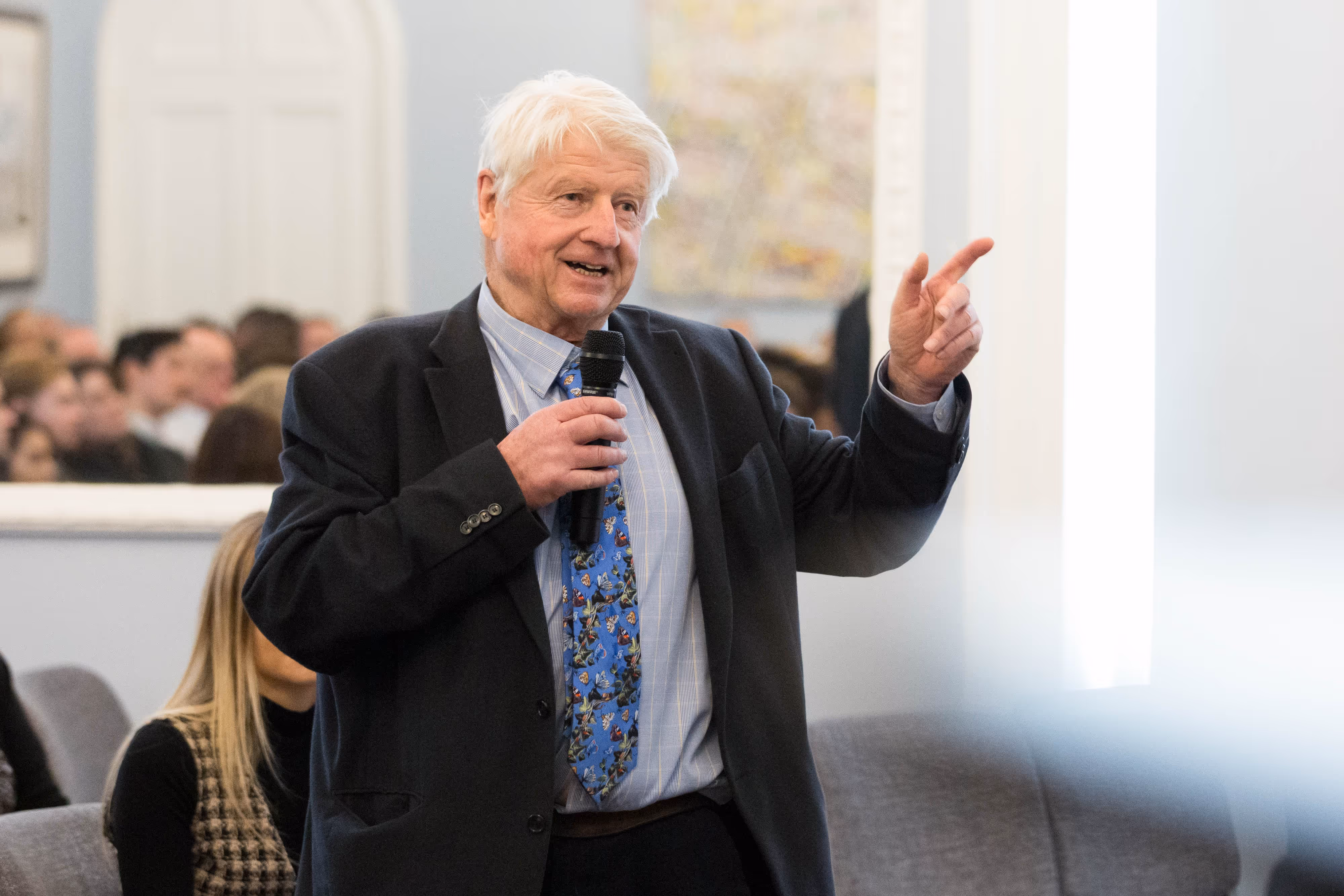 Stanley Johnson speaks into a microphone and gestures whilst standing in a room with an audience.