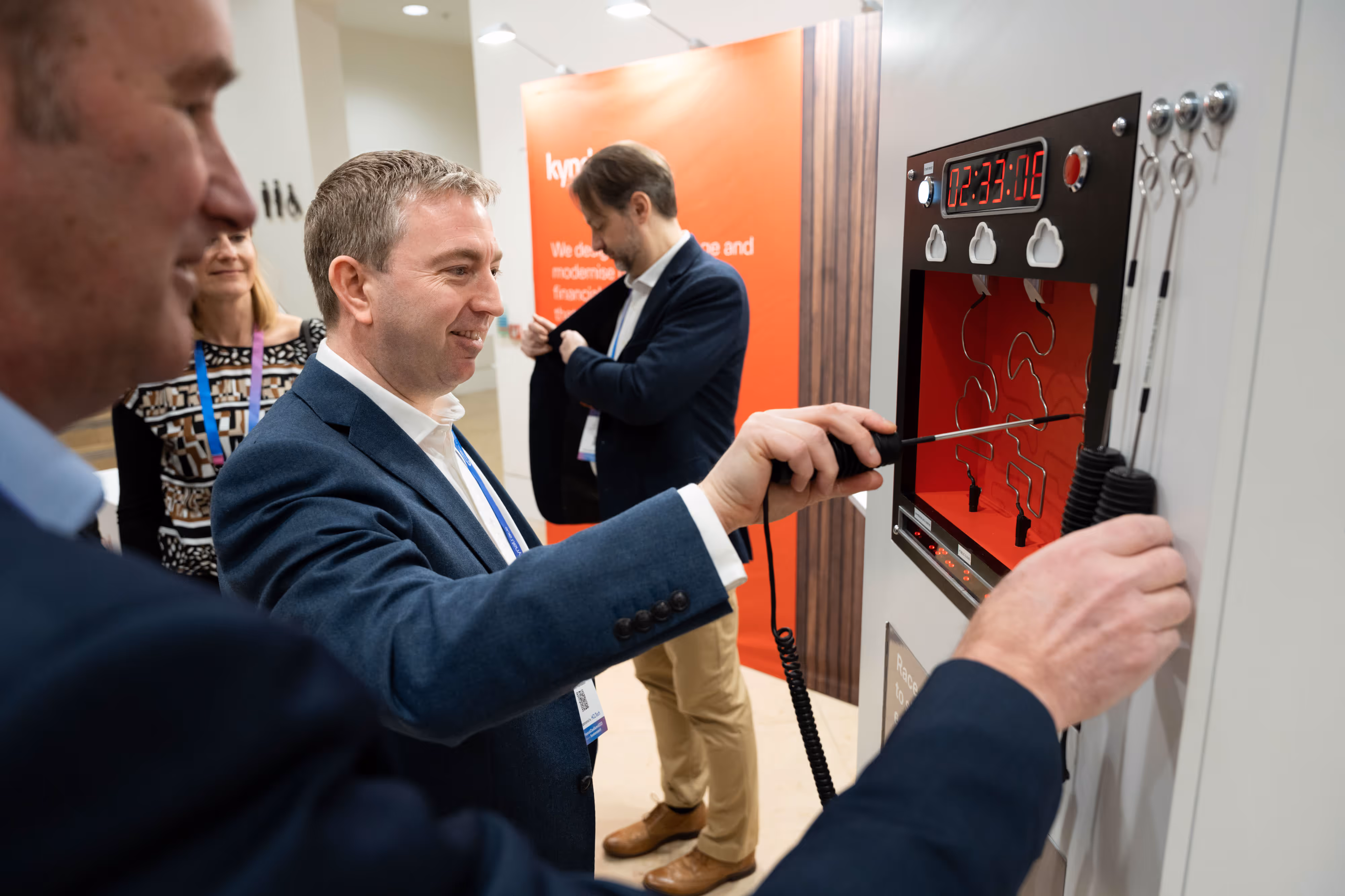 A man moves a metal rod through a wire maze game whilst others watch at an event stand.