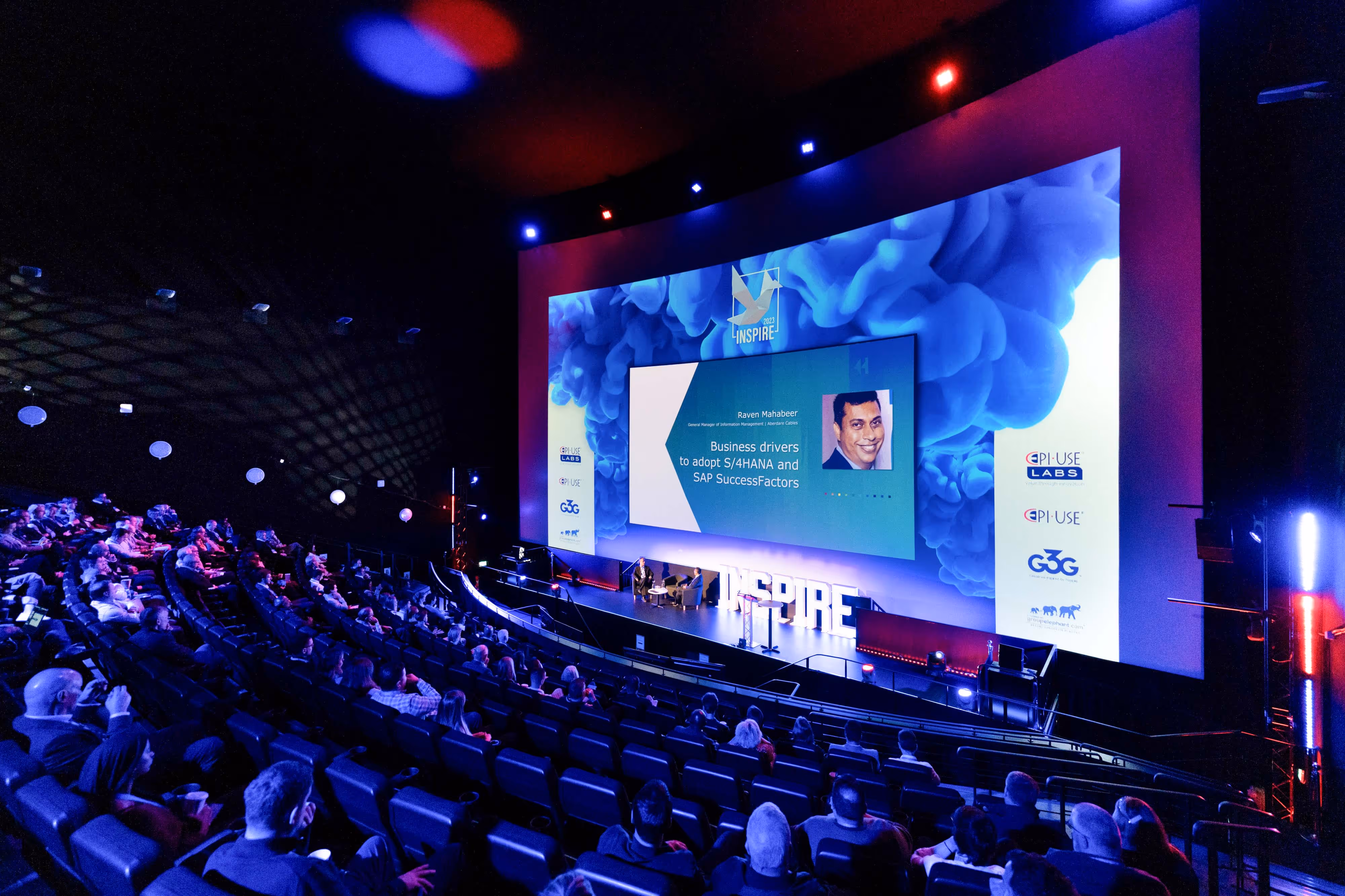 Audience watches a speaker present on a large screen at a tech conference in a modern, curved auditorium.