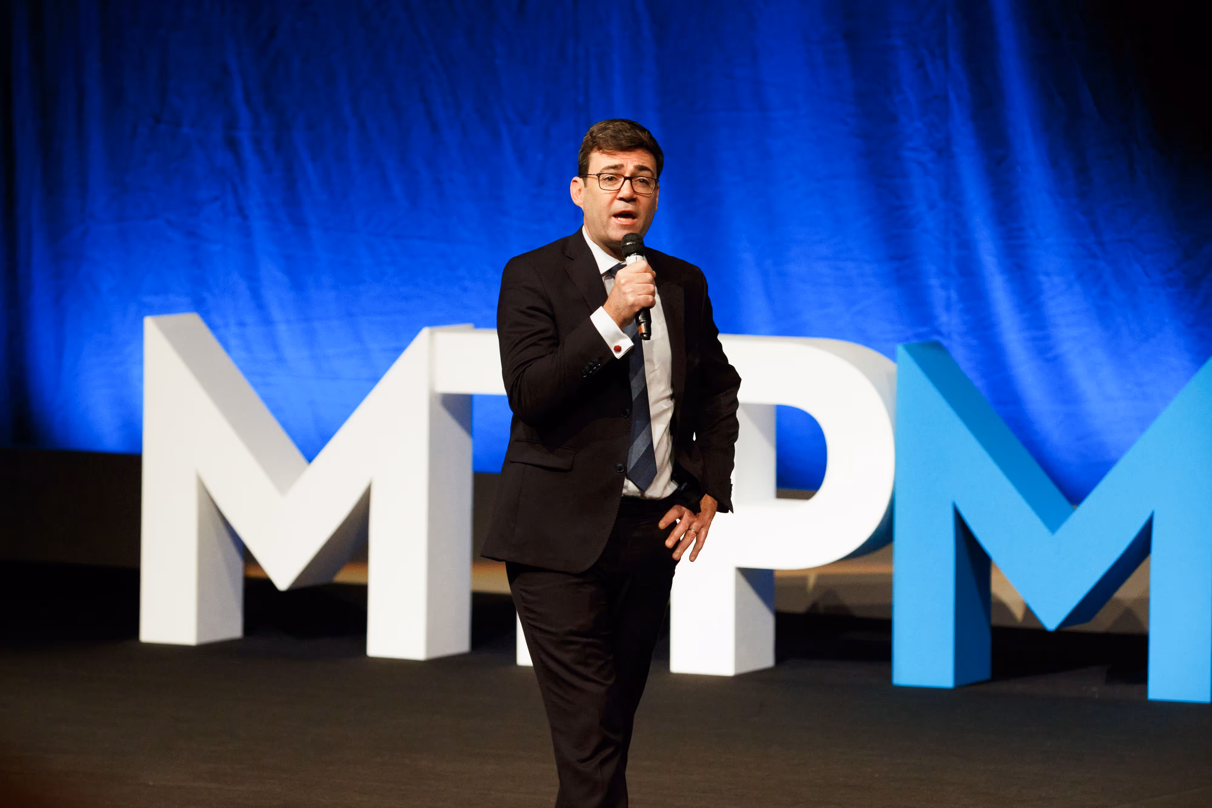 Andy Burnham speaks into a microphone on stage in front of large white and blue letters at a conference in Manchester.