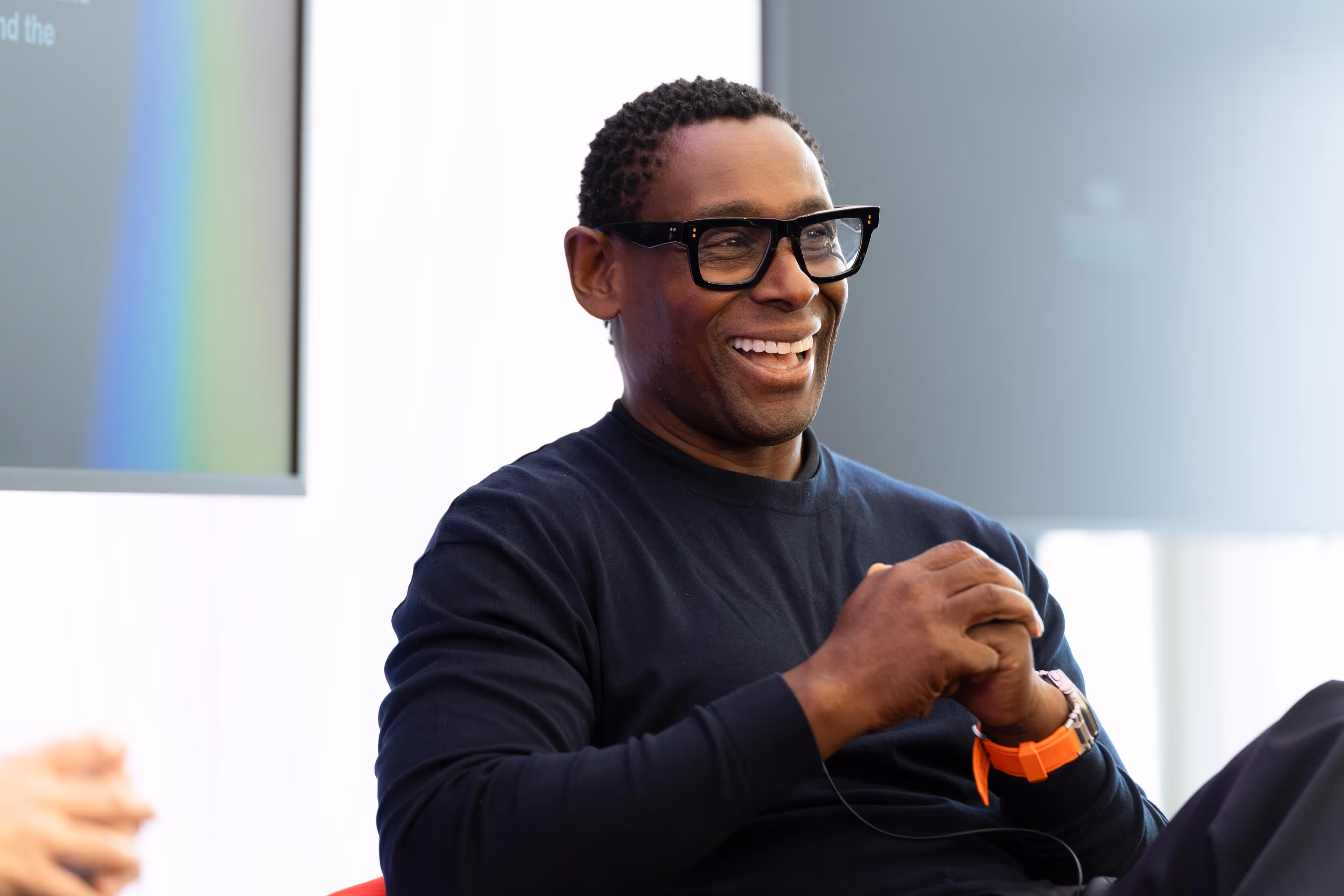 A smiling man wearing glasses and a dark jumper sits and talks, holding his hands together.