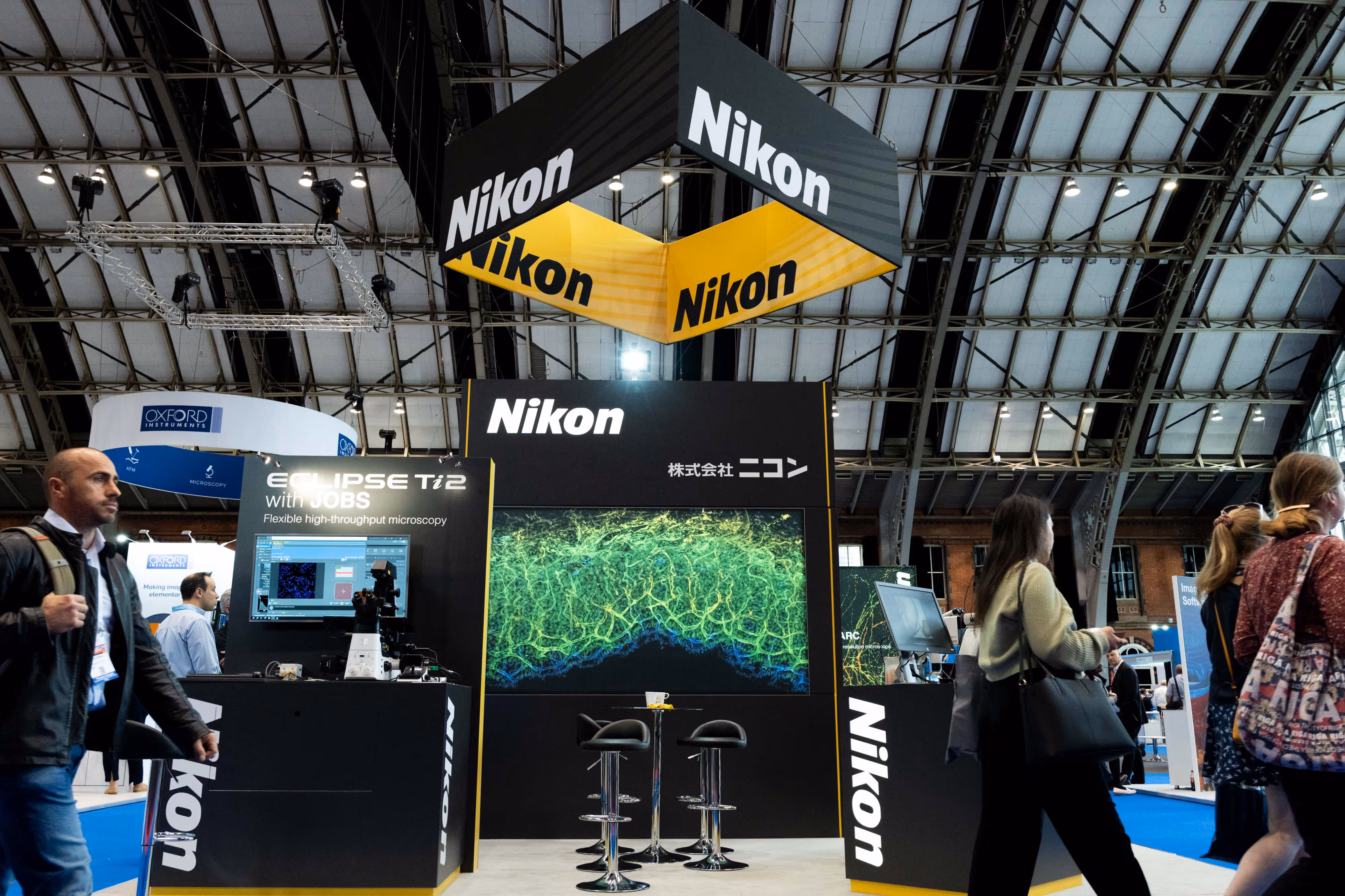 People walk past a Nikon exhibition stand with digital displays at a convention centre.