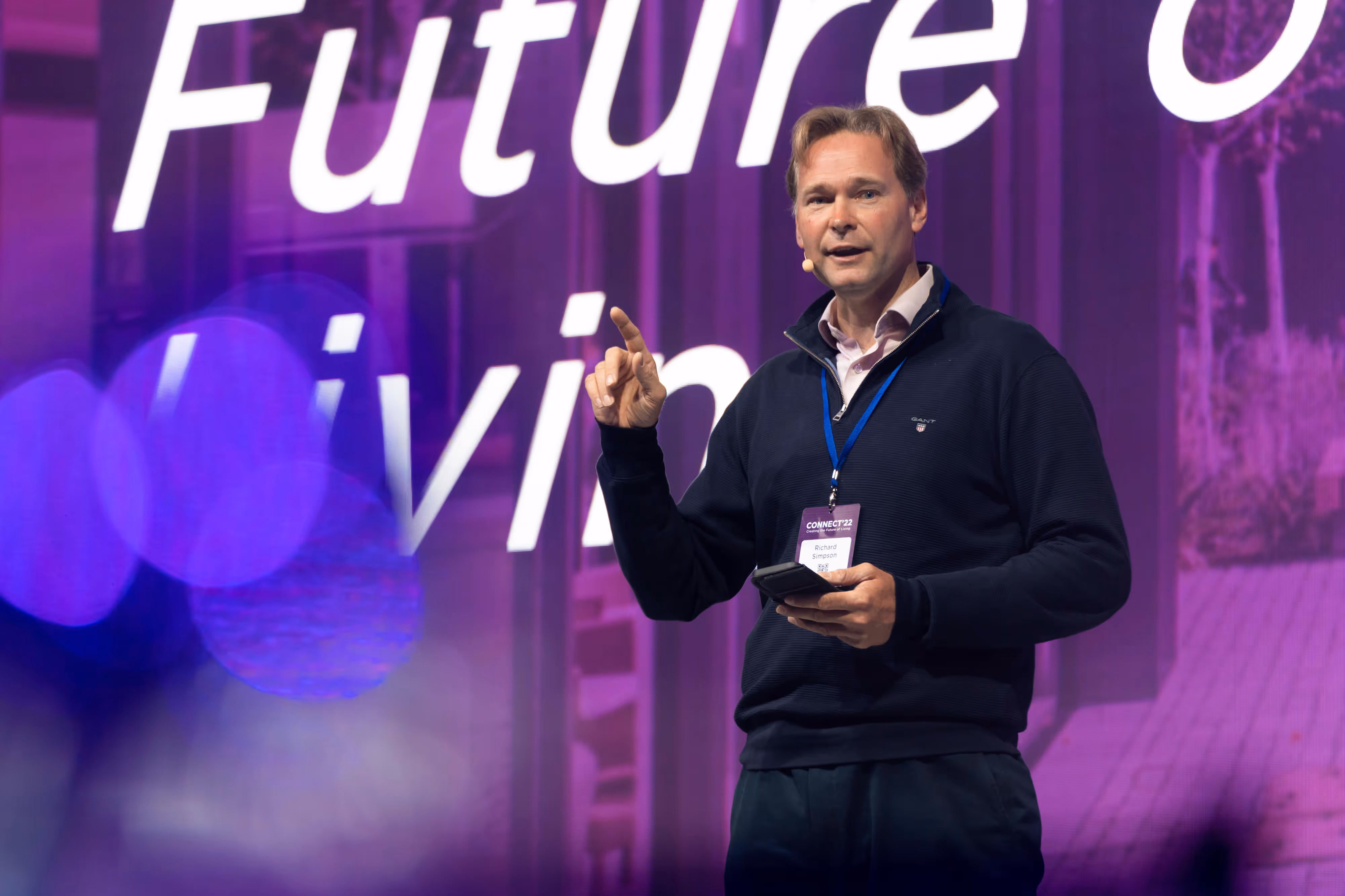 Man with microphone and badge speaks on stage in front of a purple background with large text.