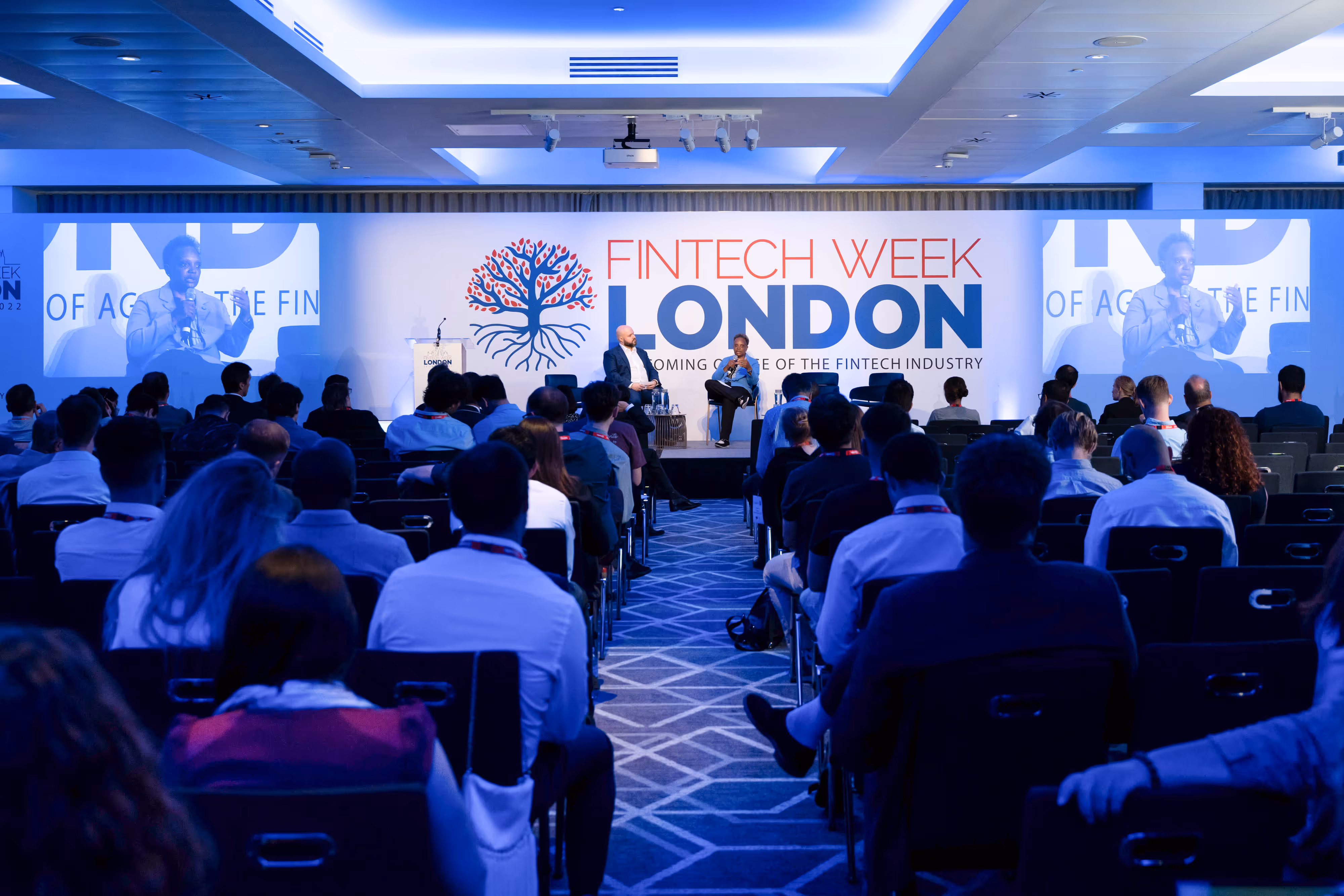 A man and woman in a fireside chat in front of a group of people in a conference room.