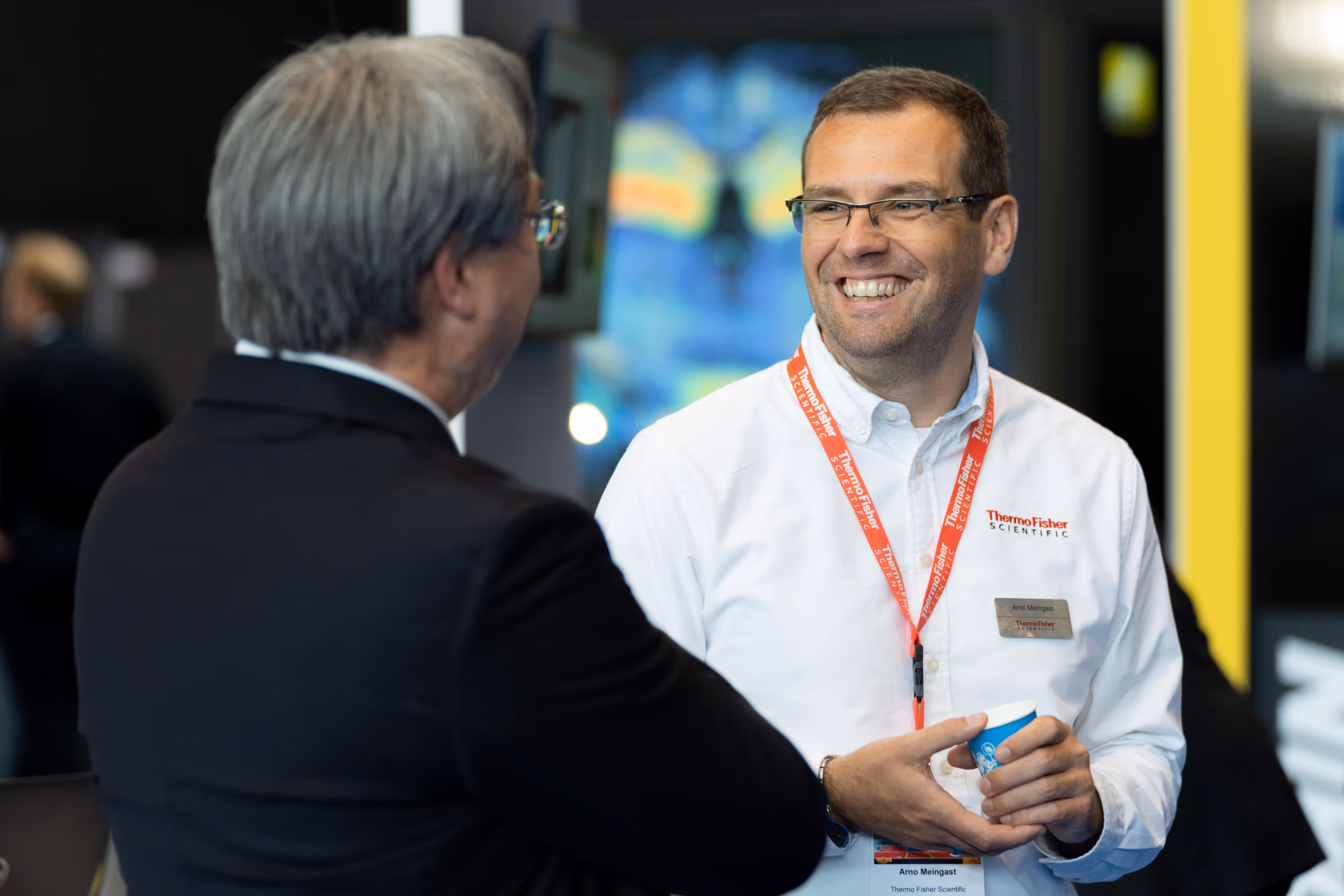 Two men in conversation at a conference; one is smiling and wearing a white shirt with a red lanyard and name badge.