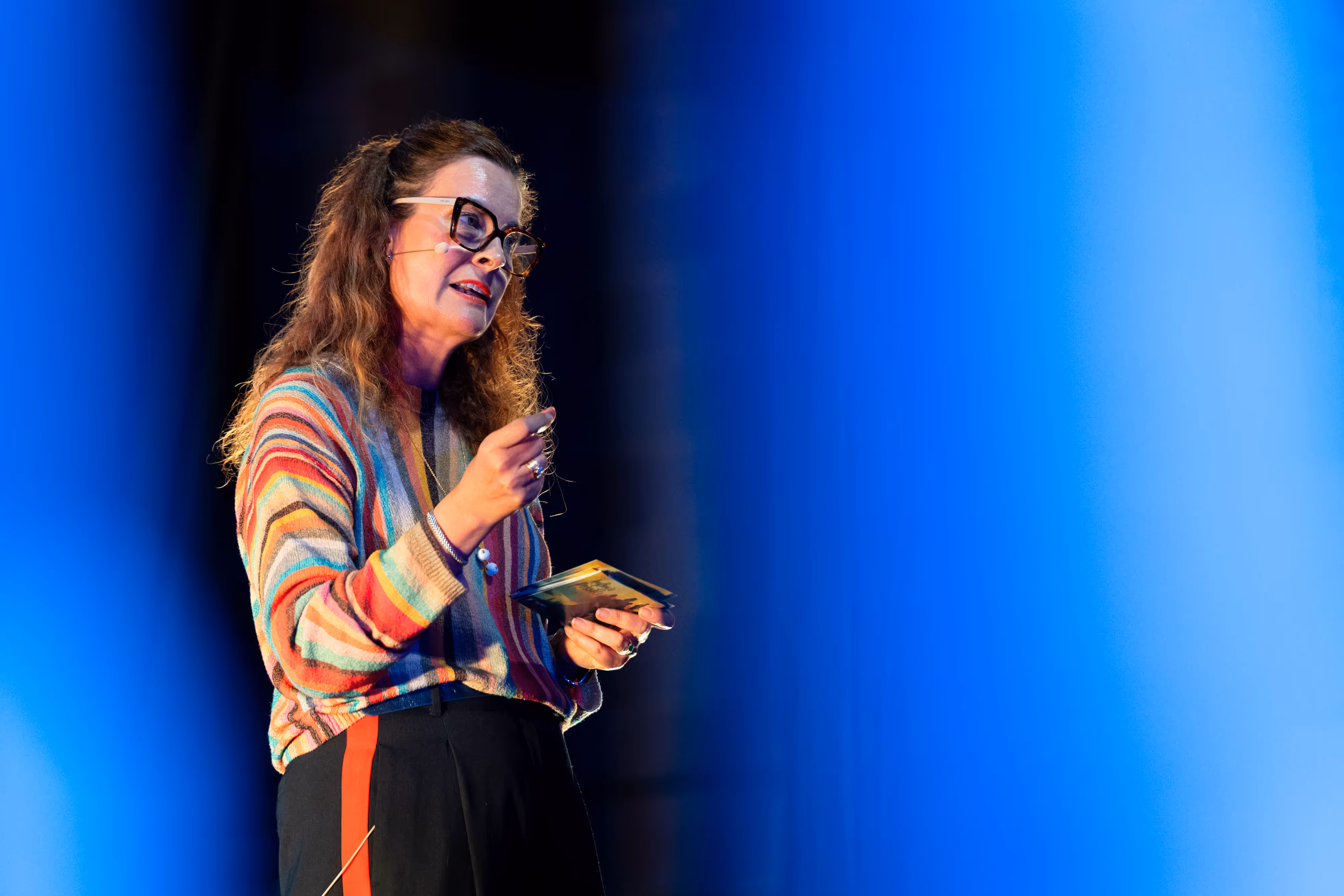 Woman in glasses and a colourful jumper speaks on stage, holding a notecard, with blue lights in the foreground.