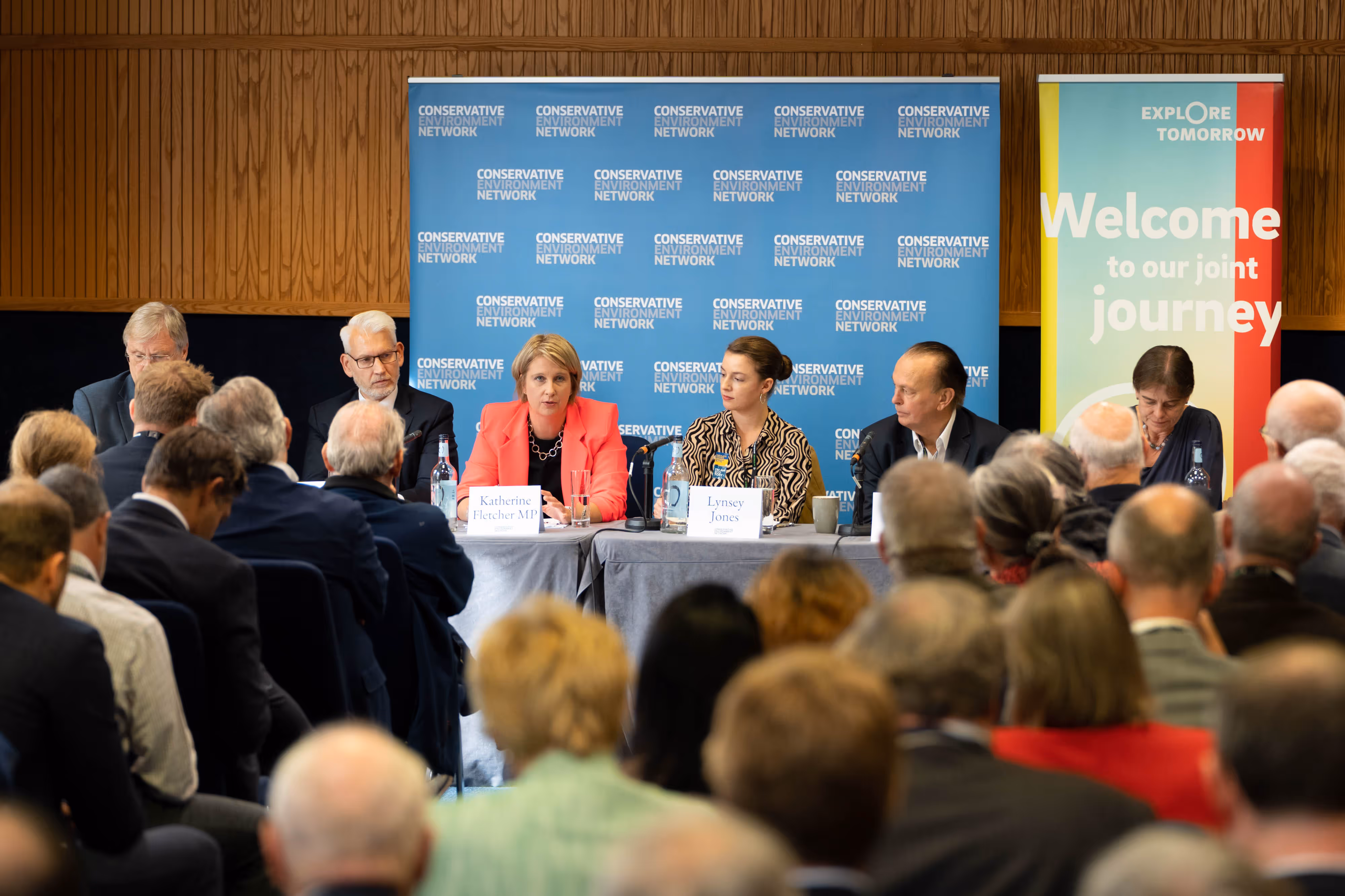 A panel of political figures speaks to an audience at an event with banners and microphones on the table.