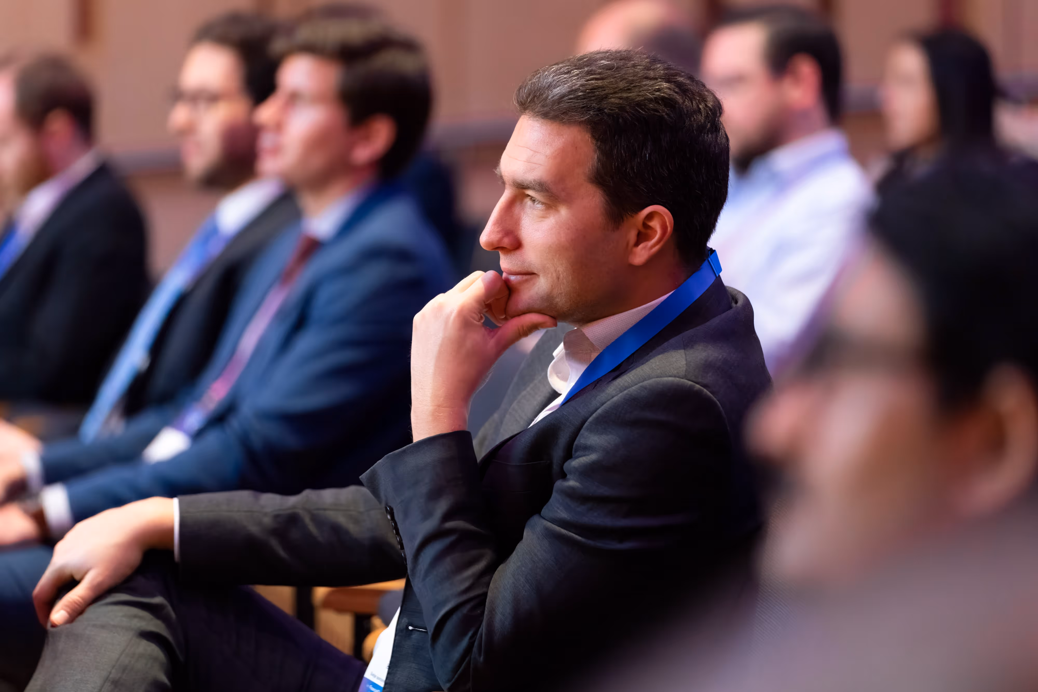 Man in a suit attentively listens at a conference, surrounded by other seated attendees in business attire.