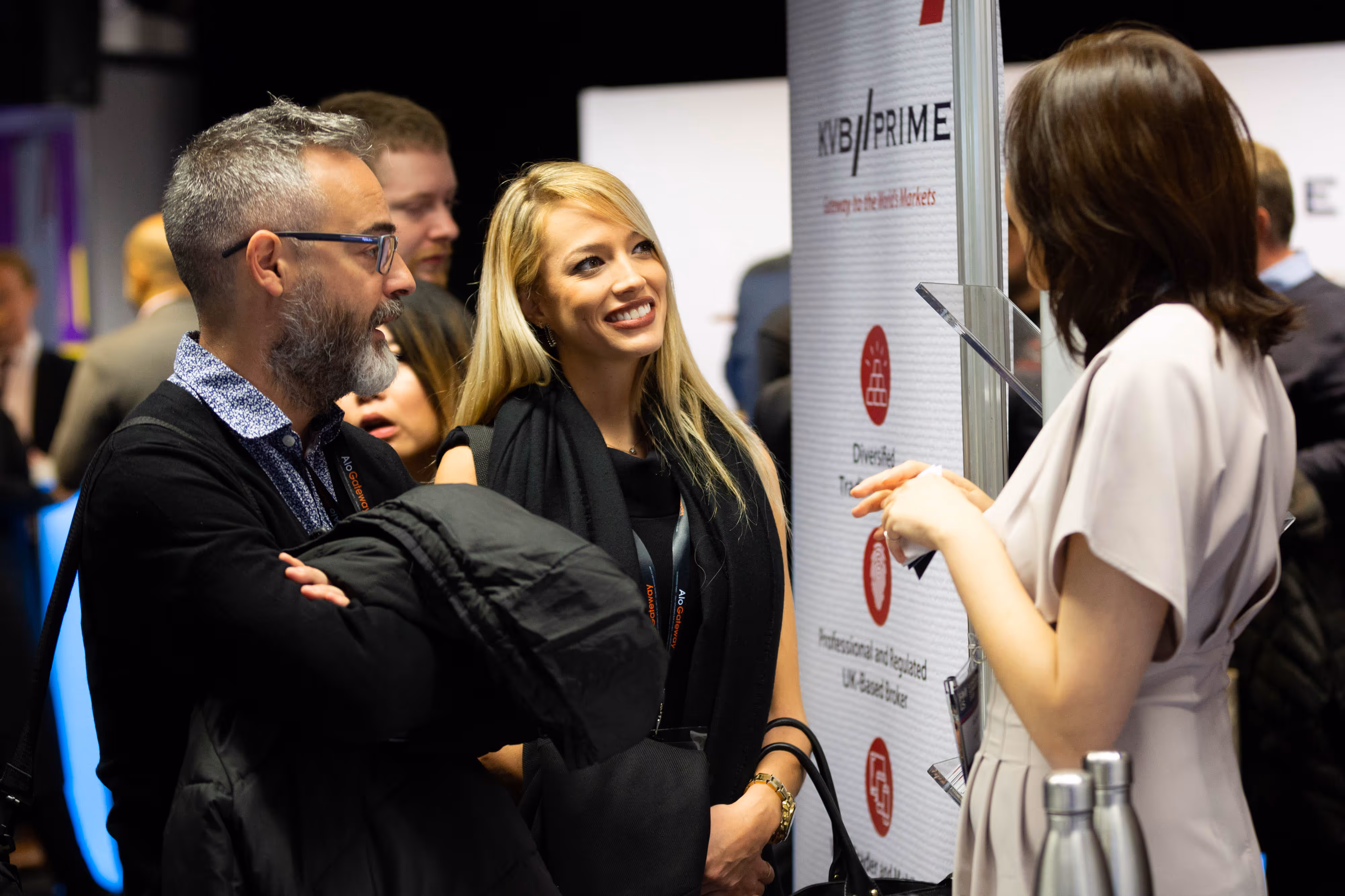 Three people stand and converse at a business event, smiling near a display board with information and logos.