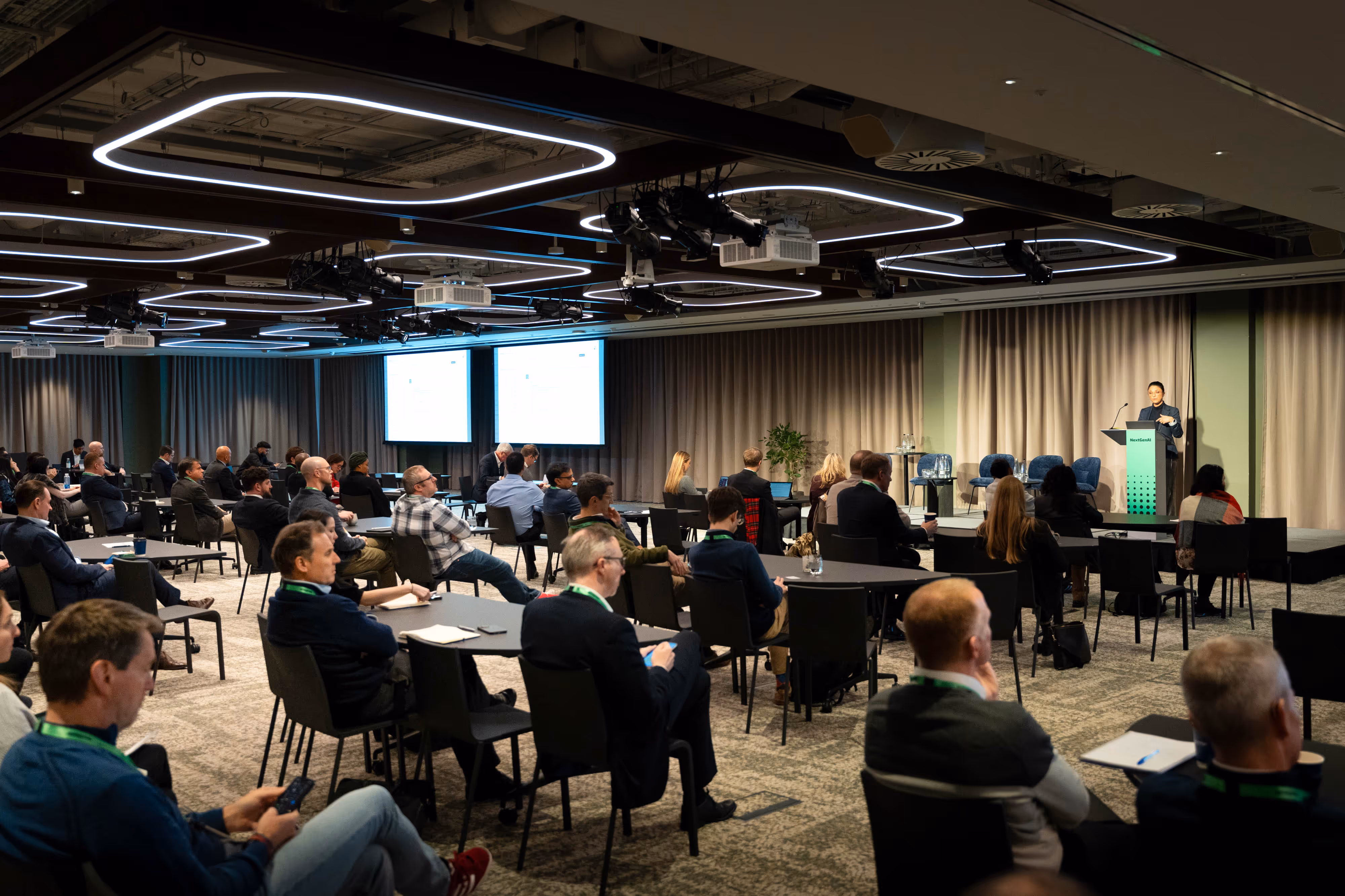 Conference attendees sit at tables, listening to a speaker on stage with presentation screens in a modern meeting room.