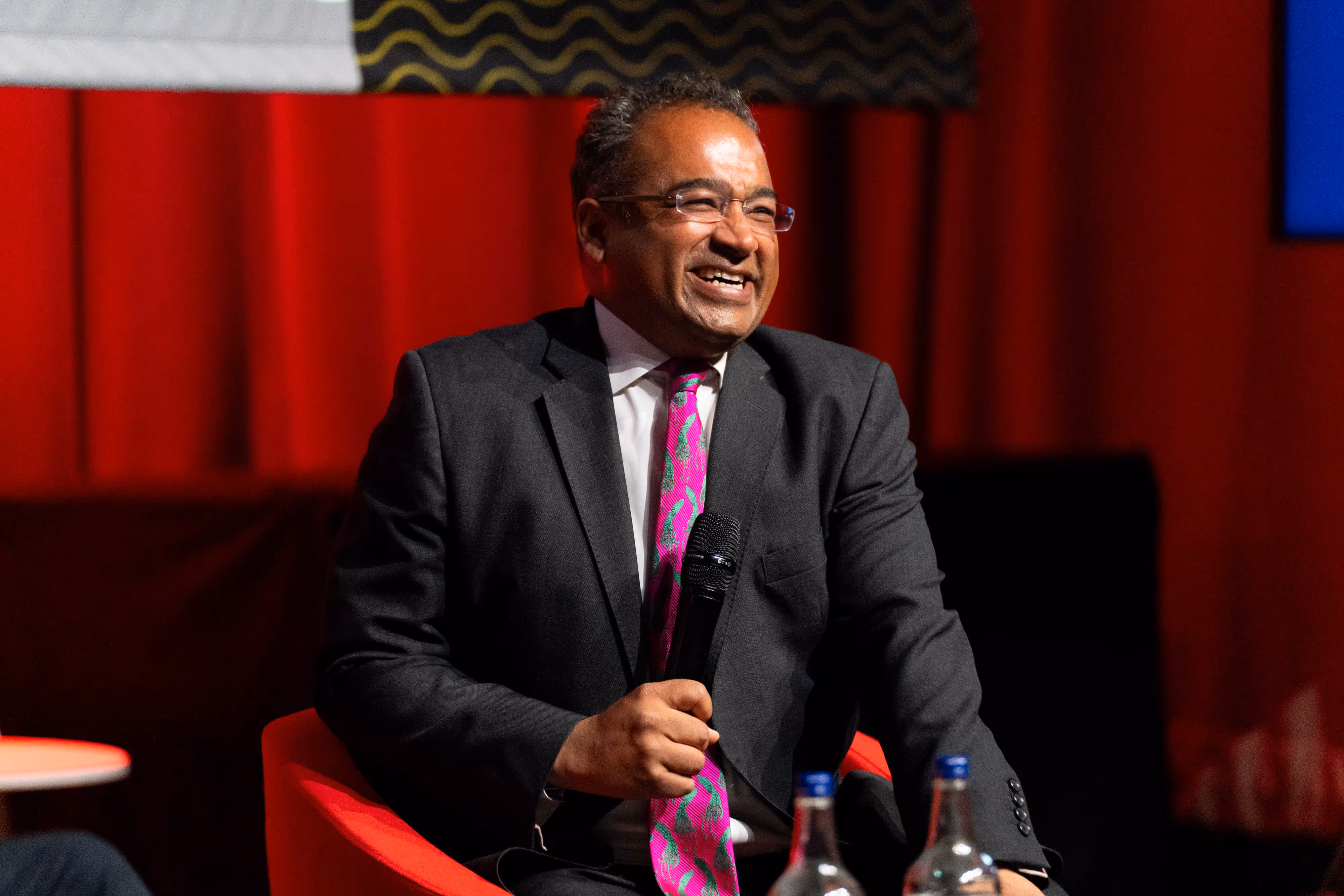 A celebrity journalist in a suit and pink tie sits on a red chair, smiling and holding a microphone on stage.
