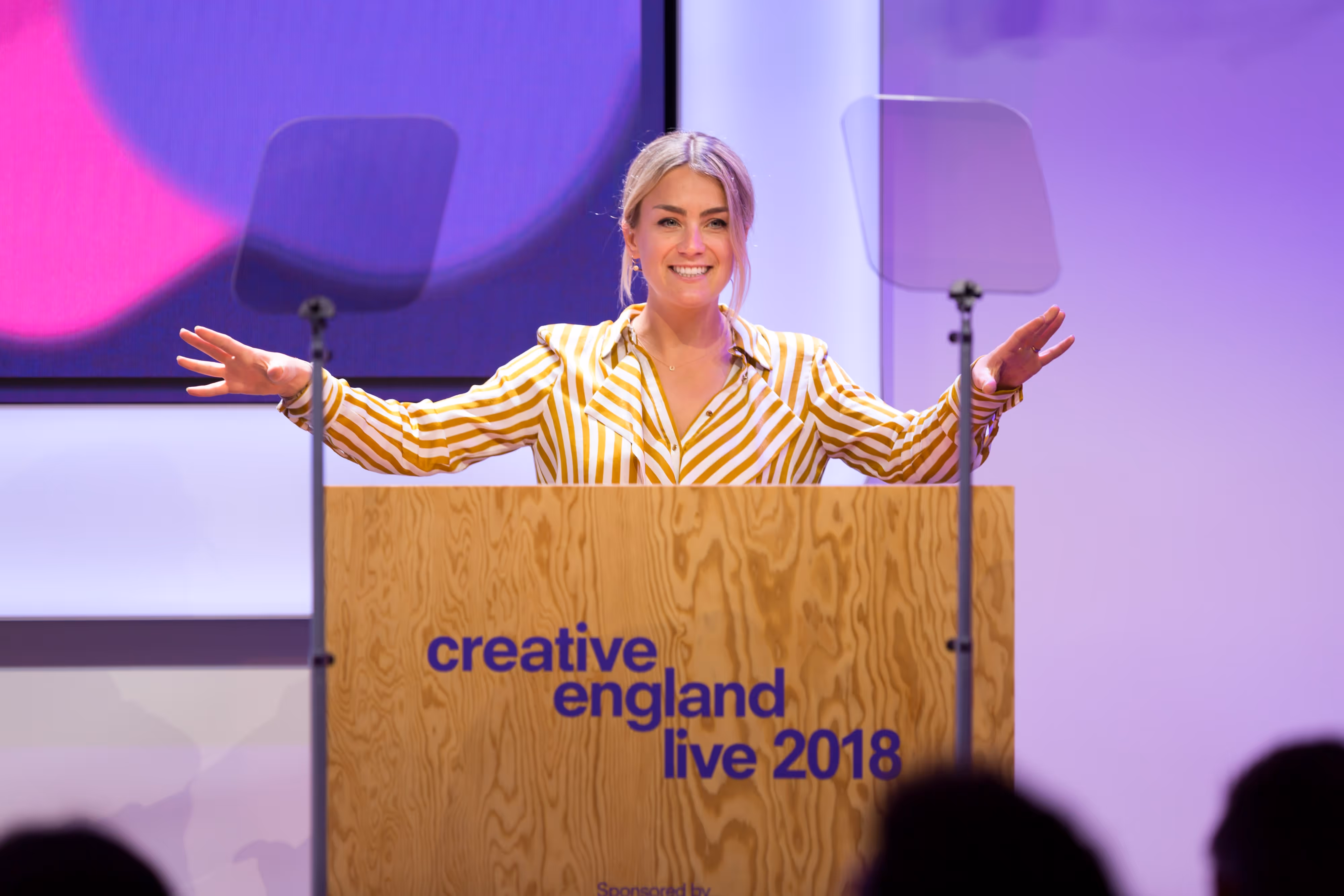 A smiling woman stands at a podium labelled "creative england live," speaking at an event.