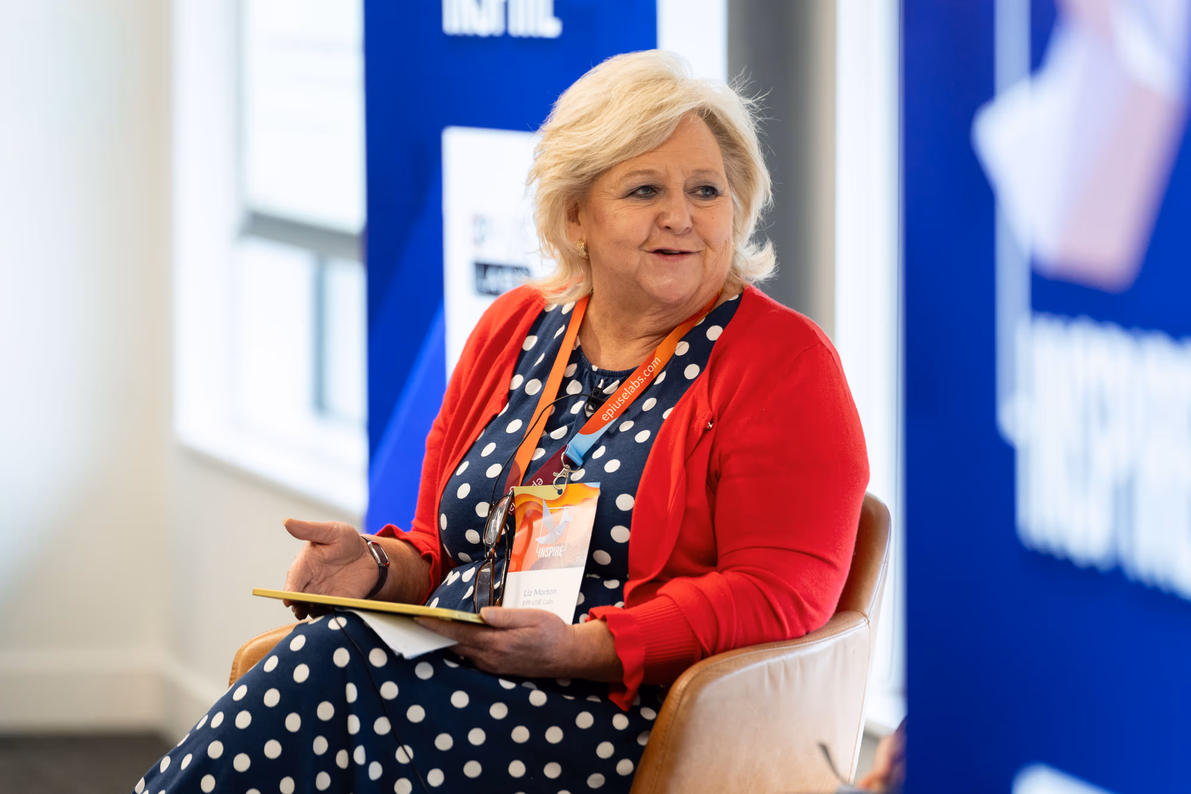A woman in a red cardigan and polka dot dress speaks whilst seated, holding a notepad at an etc venues event.