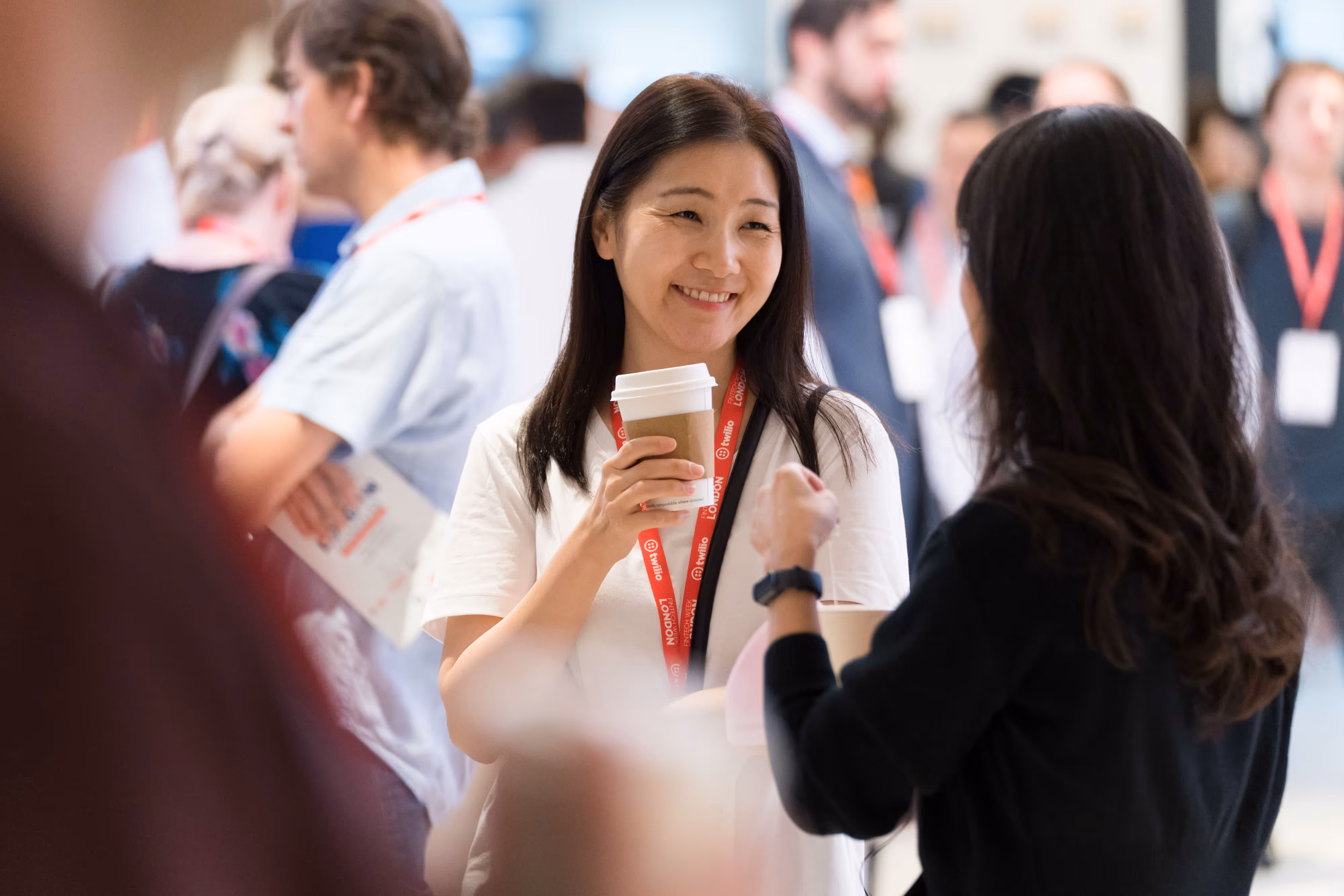 Smiling woman holding a coffee cup talks with another person at a busy conference or networking event.