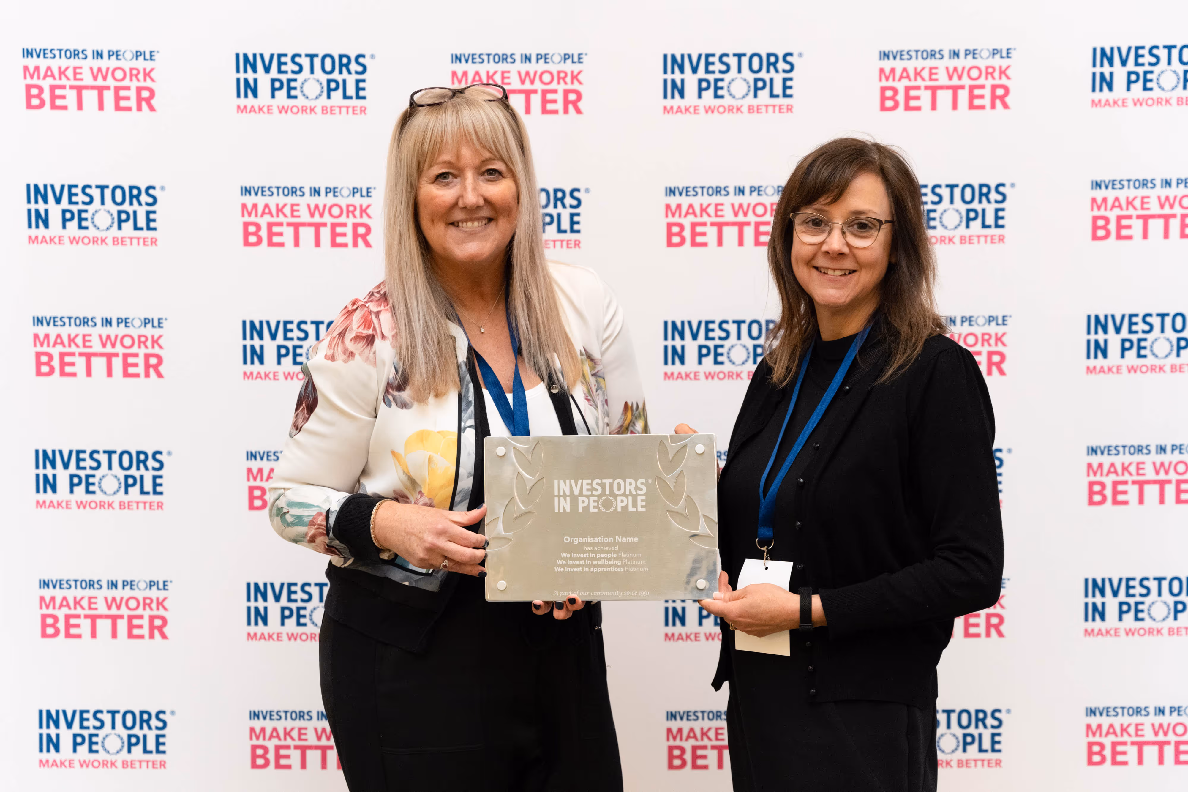 Two women holding an Investors in People award plaque, standing in front of a branded backdrop.