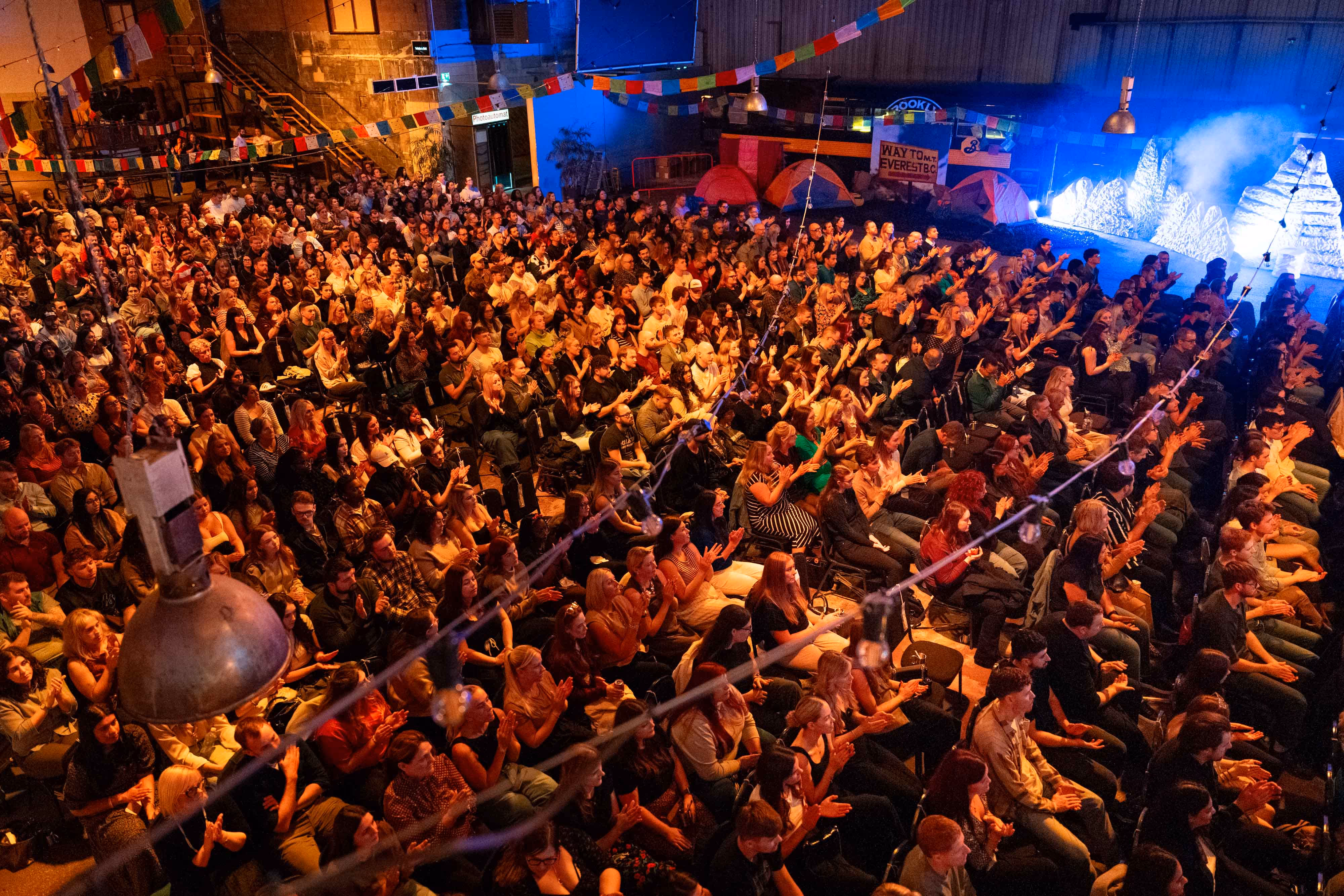A large audience sits in rows at an indoor event, watching a brightly lit stage with decorations.