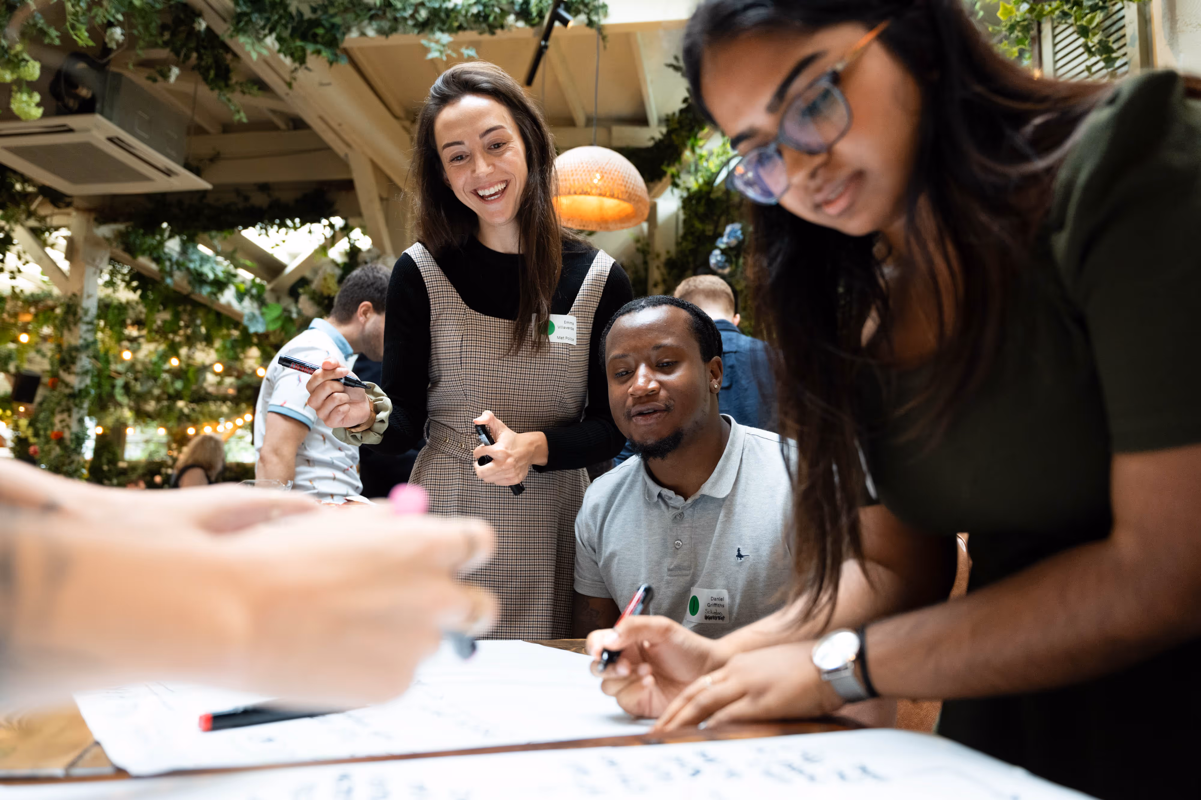 Three people collaborate and write on a large sheet of paper in a bright, plant-filled indoor space.