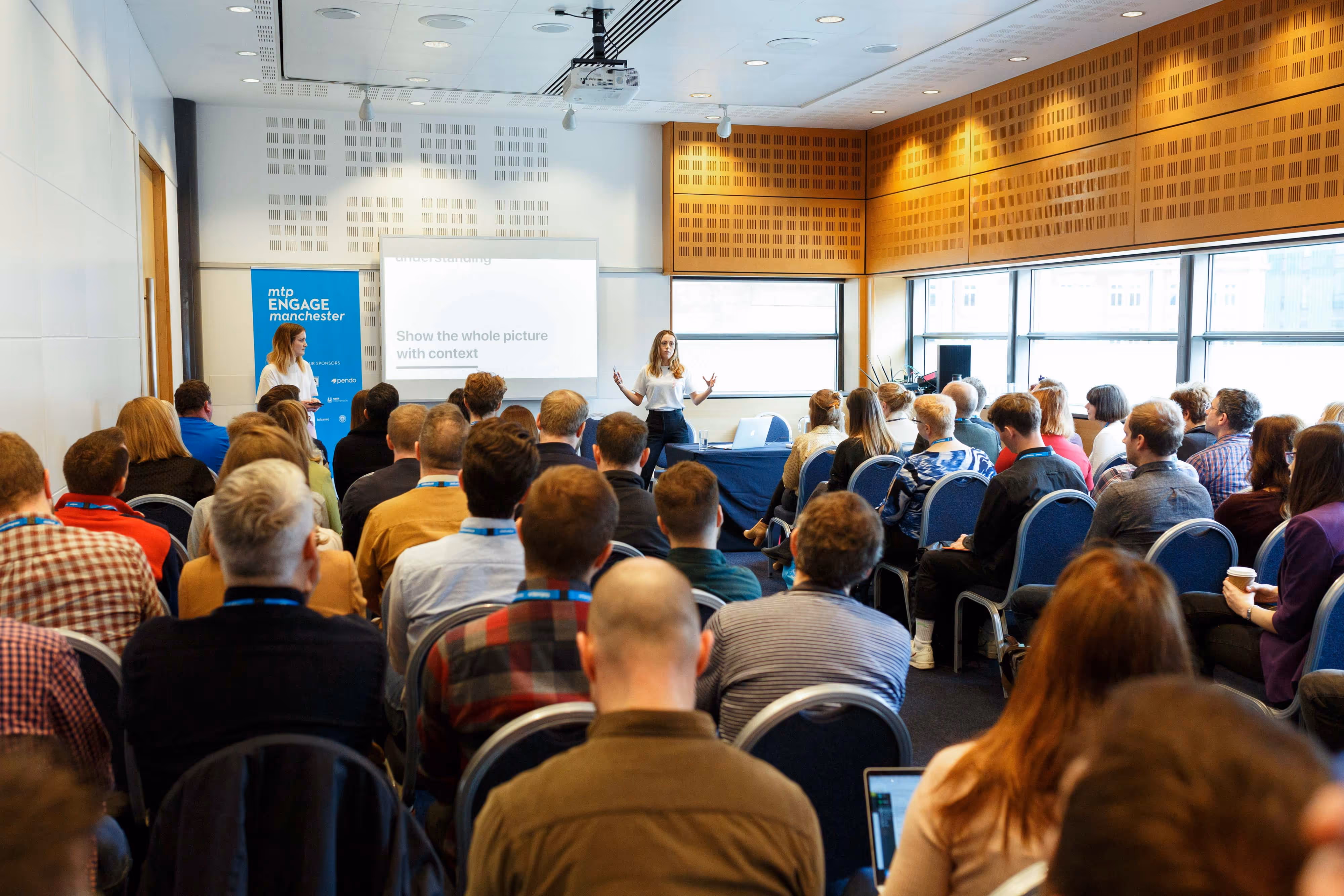 A woman gives a presentation to a seated audience in a bright, modern conference room.