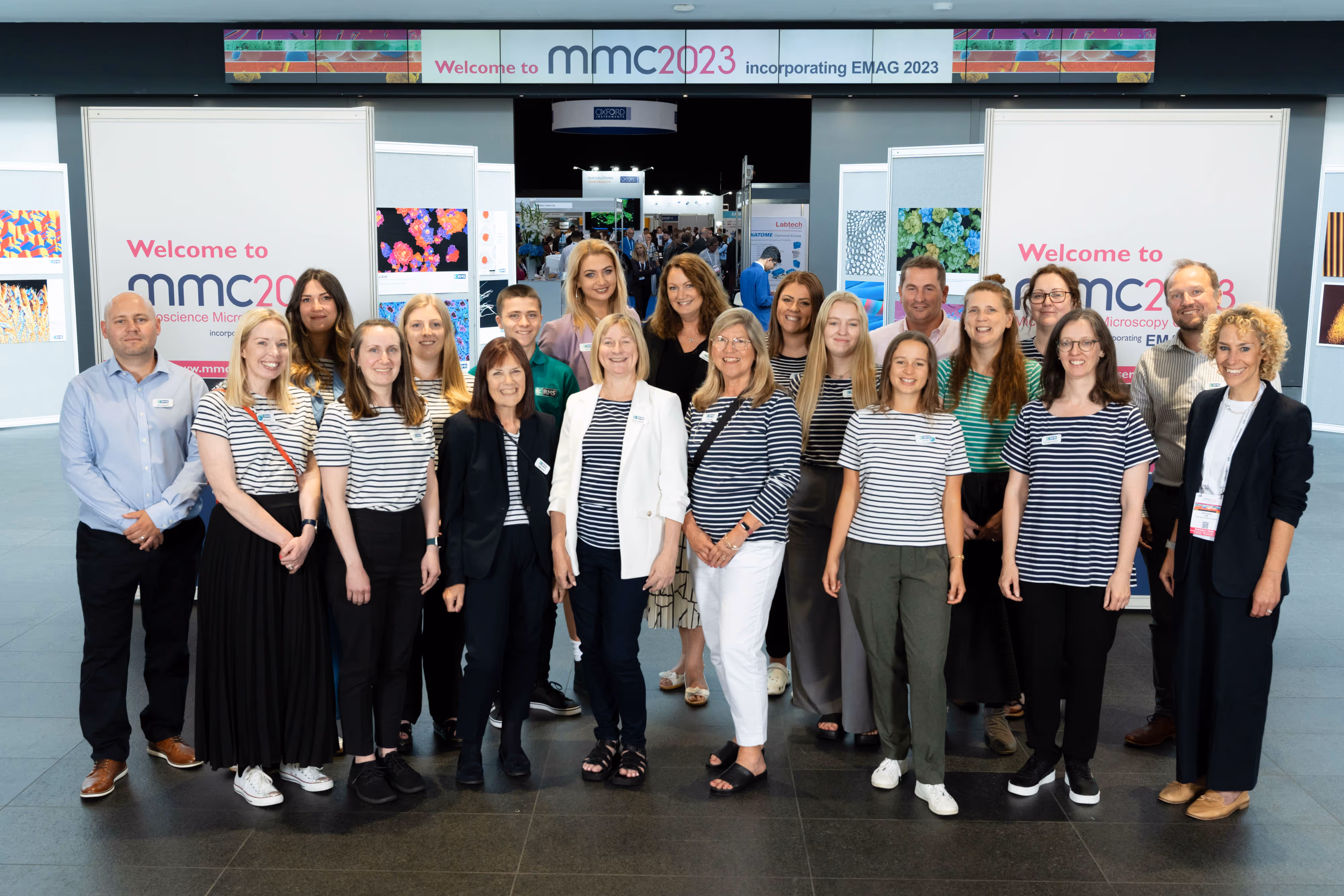 A group of people pose and smile together for a team photo at the entrance of the MMC 2023 conference.