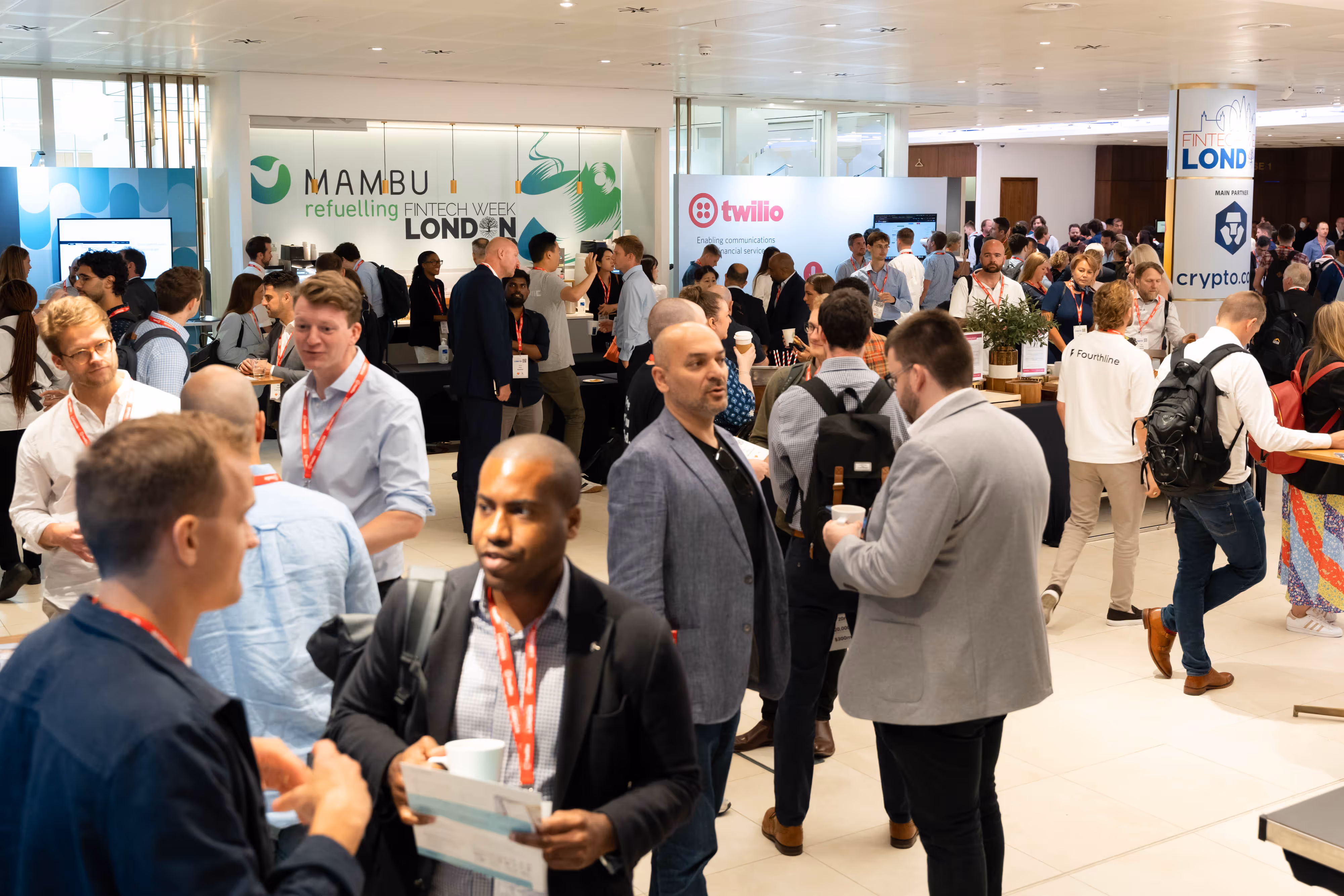 A crowded conference hall with people networking; sponsor stands for Mambu and Twilio visible in the background.