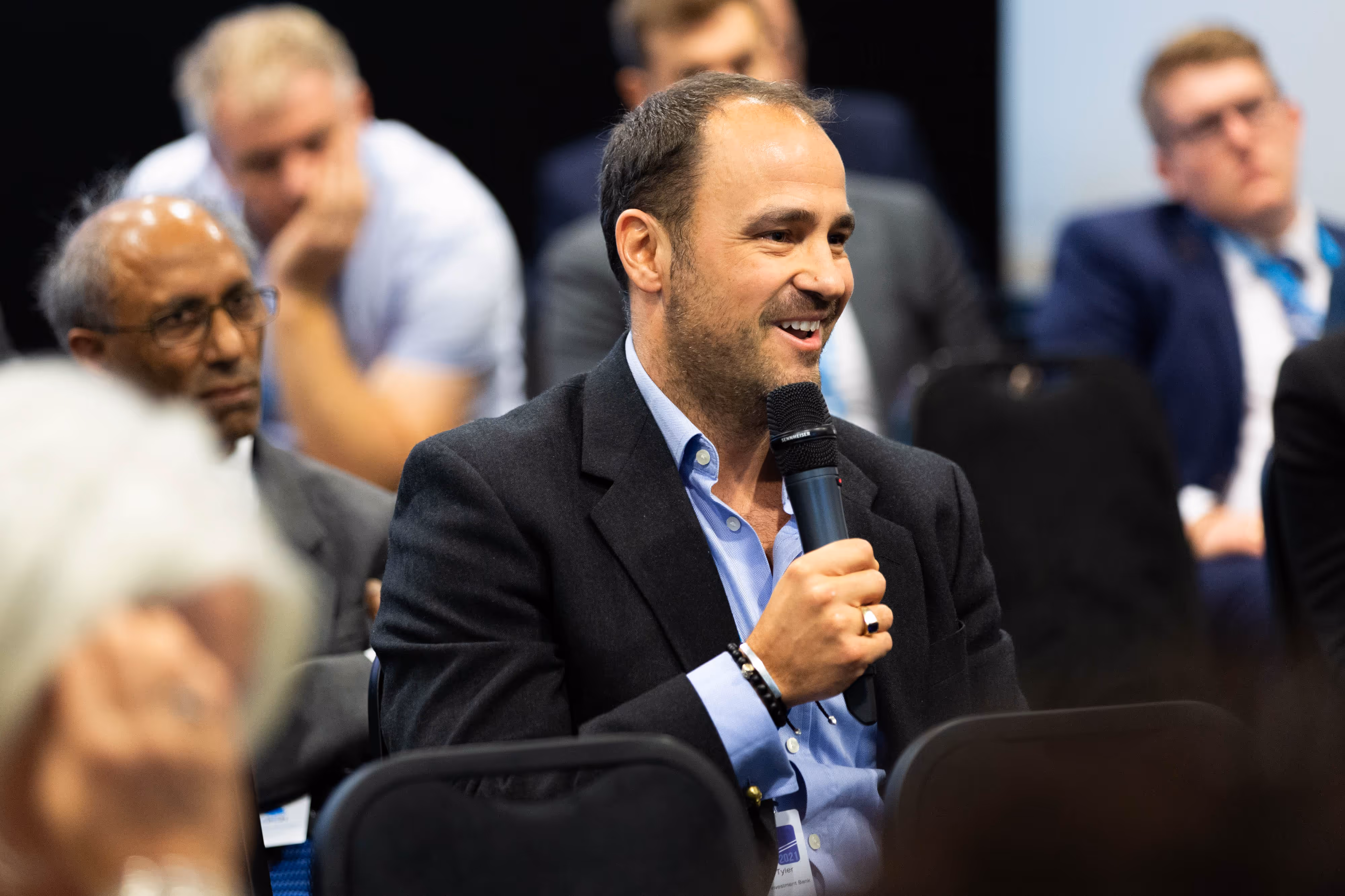 Man in a suit smiling and speaking into a microphone during a seated event, with others listening in the background.