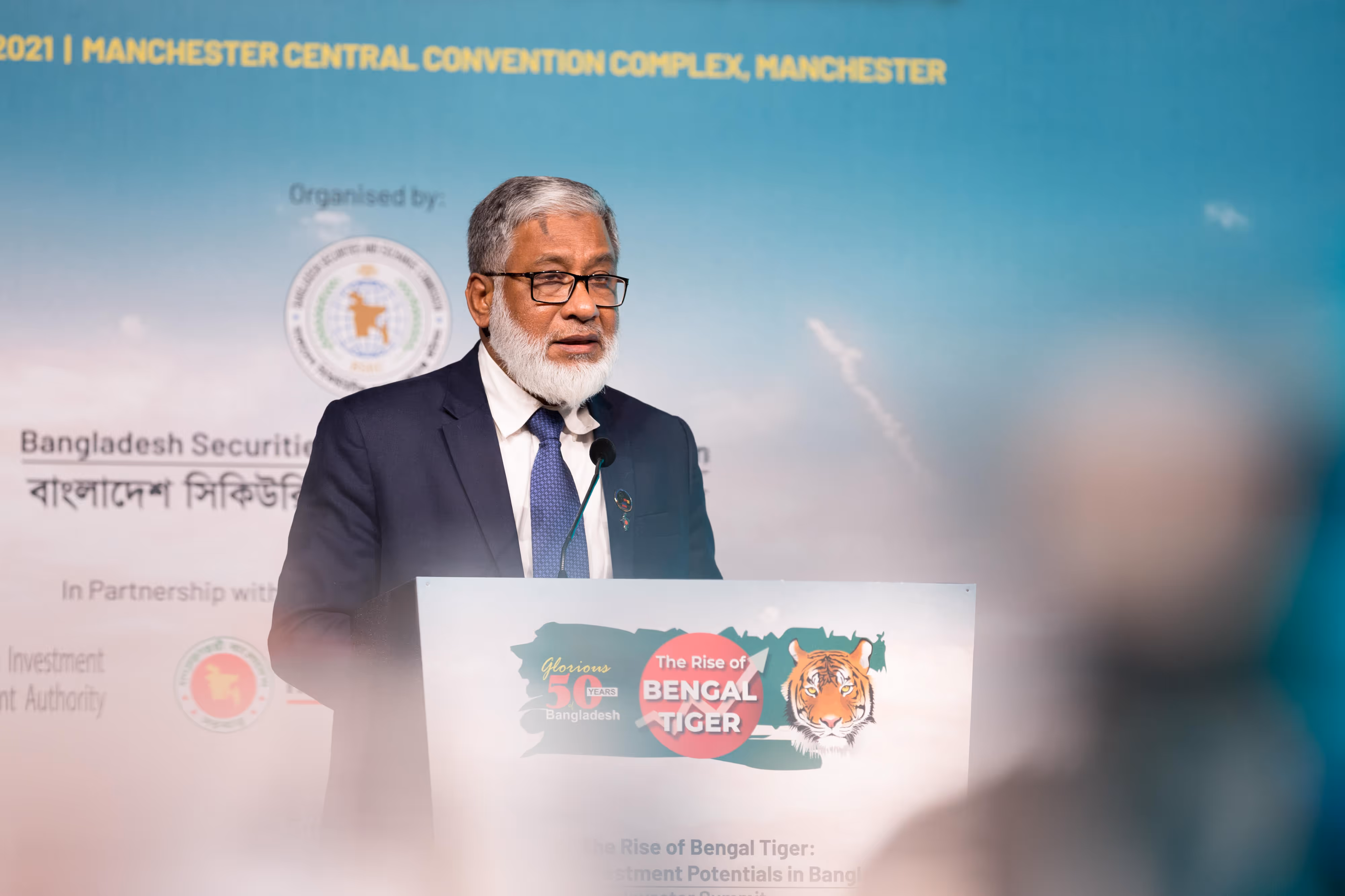 An older man in a suit speaks at a lectern during an event about investment in Bangladesh.