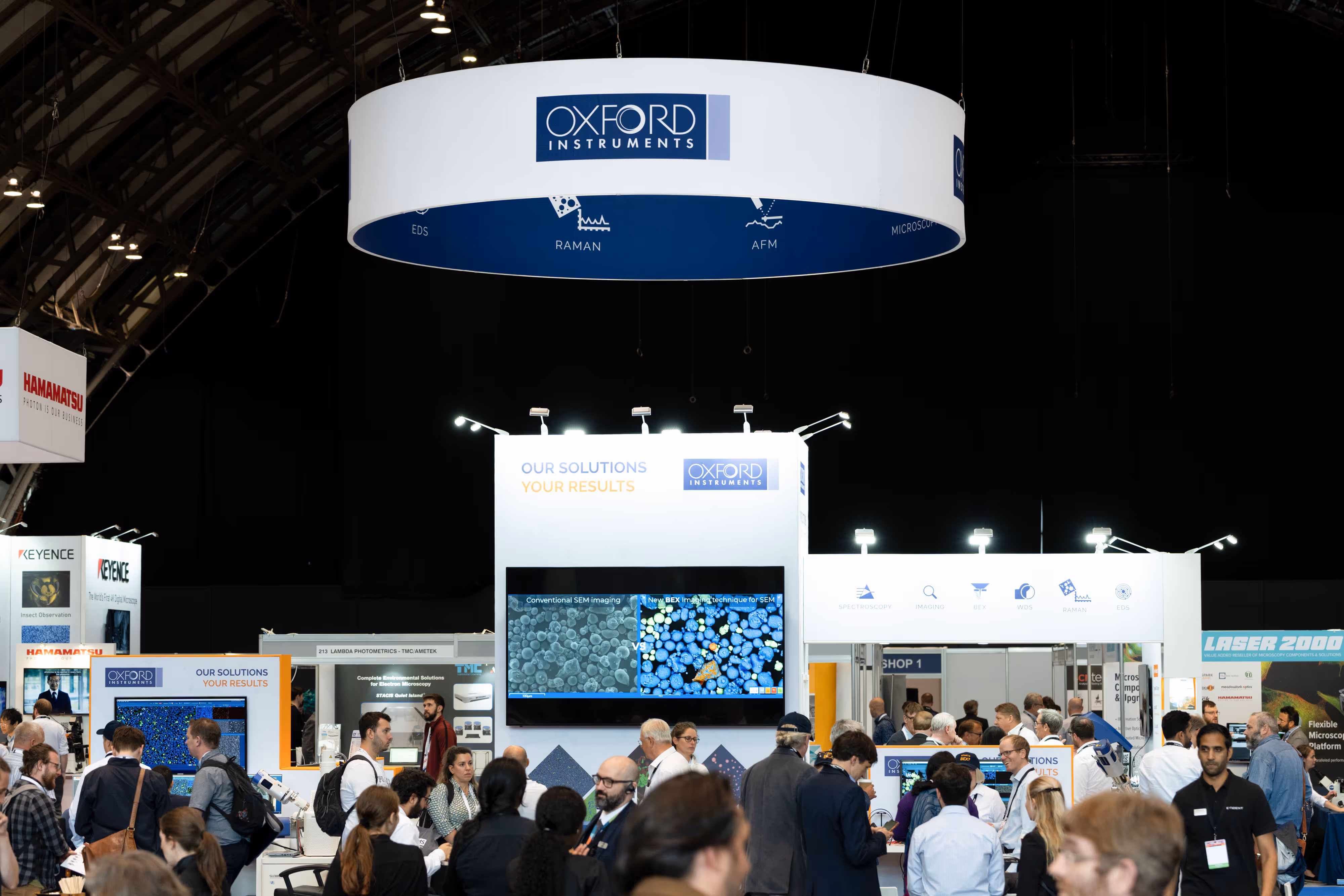 Crowd at a science and technology exhibition, featuring an Oxford Instruments stand and a large circular hanging sign.A crowded conference hall with people networking; sponsor stands for Mambu and Twilio visible in the background.