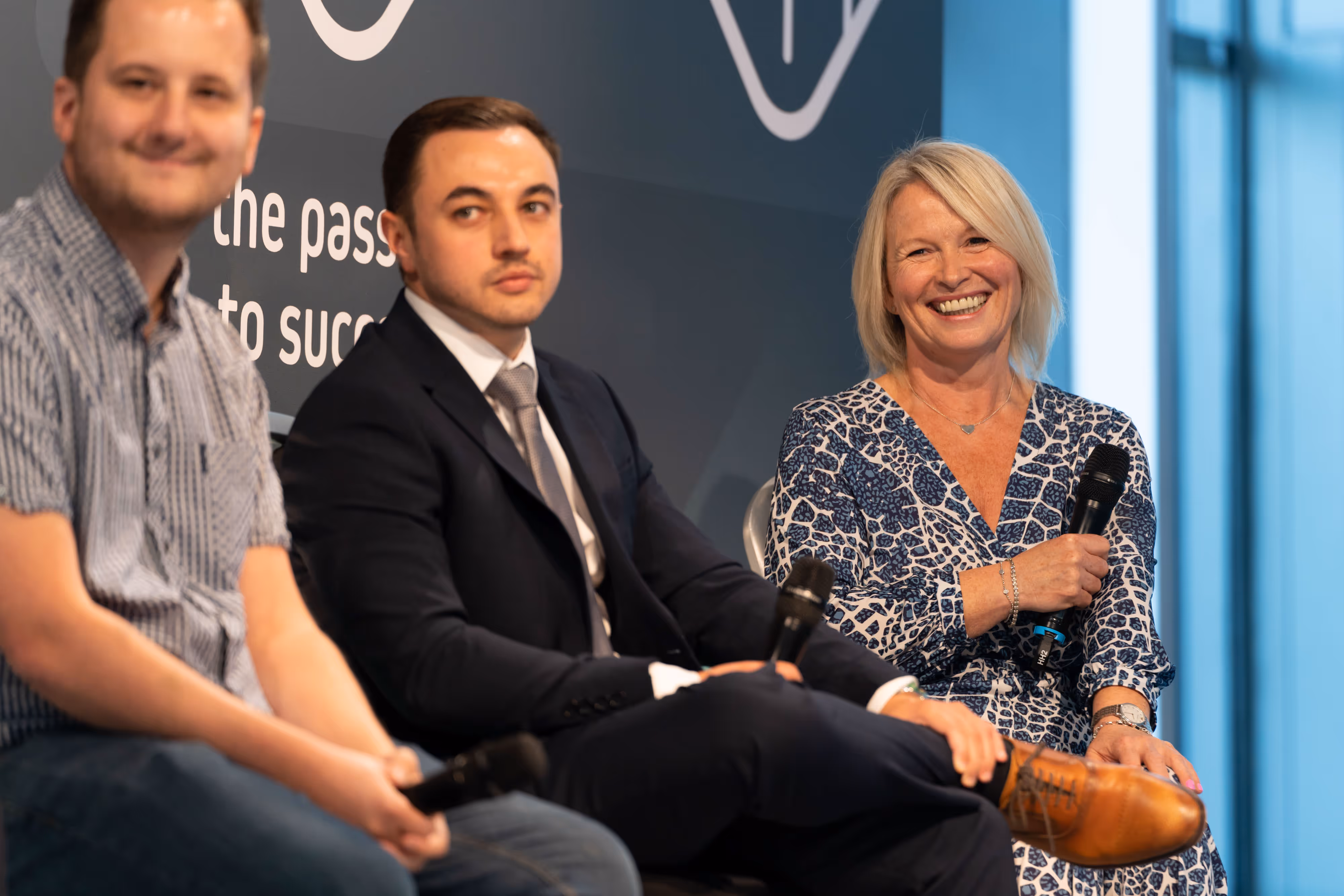 Three people sit on a panel holding microphones; one woman is smiling, and two men look towards the audience.
