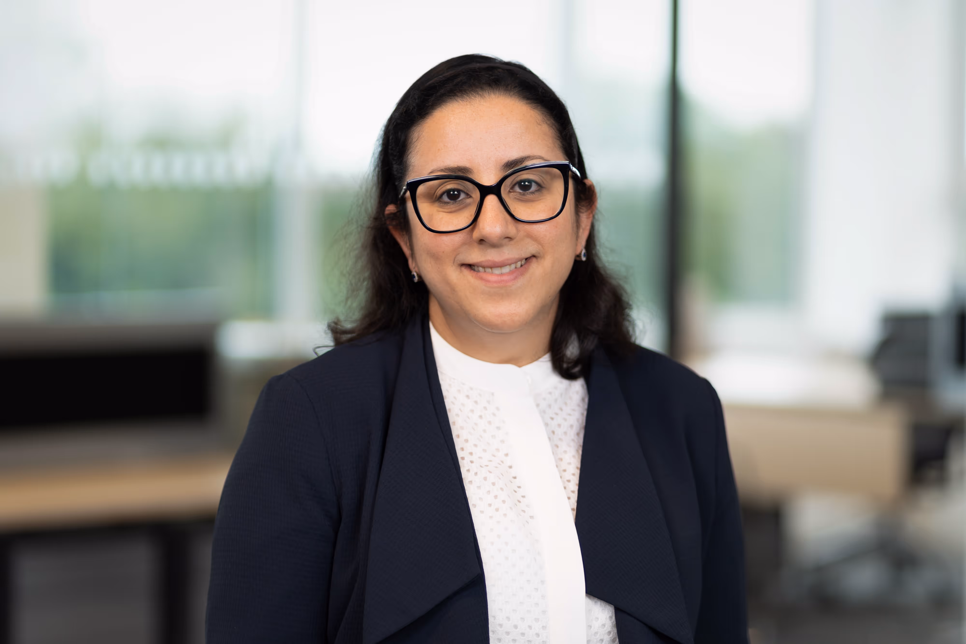 Woman with dark hair and glasses smiling, wearing a navy blazer and white blouse, in a bright office setting.