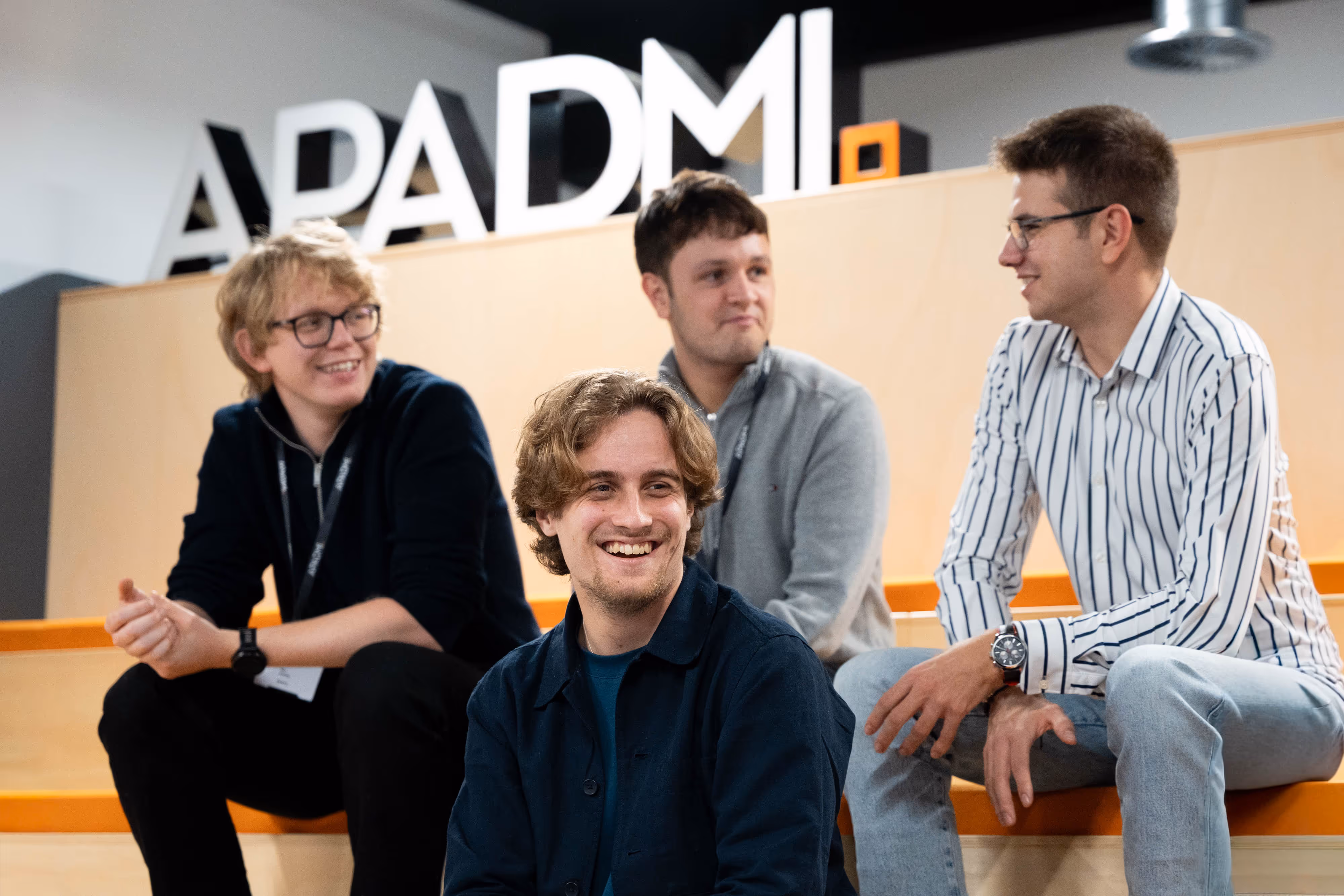 Four young men sit and chat on wooden benches with a large "APADMI" sign in the background.