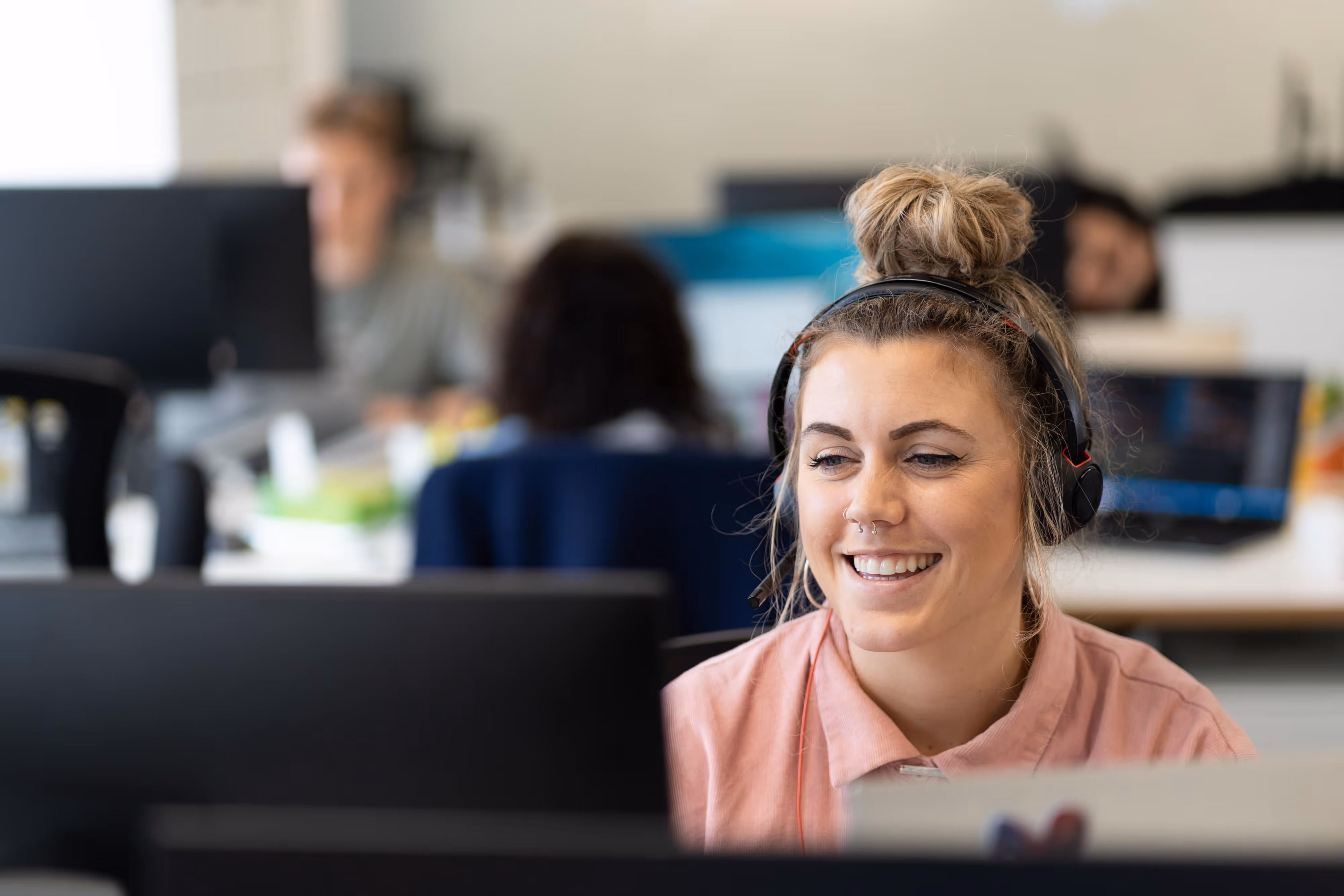 Smiling tech marketing specialist wearing headphones works at a computer in a busy office with colleagues in the background.