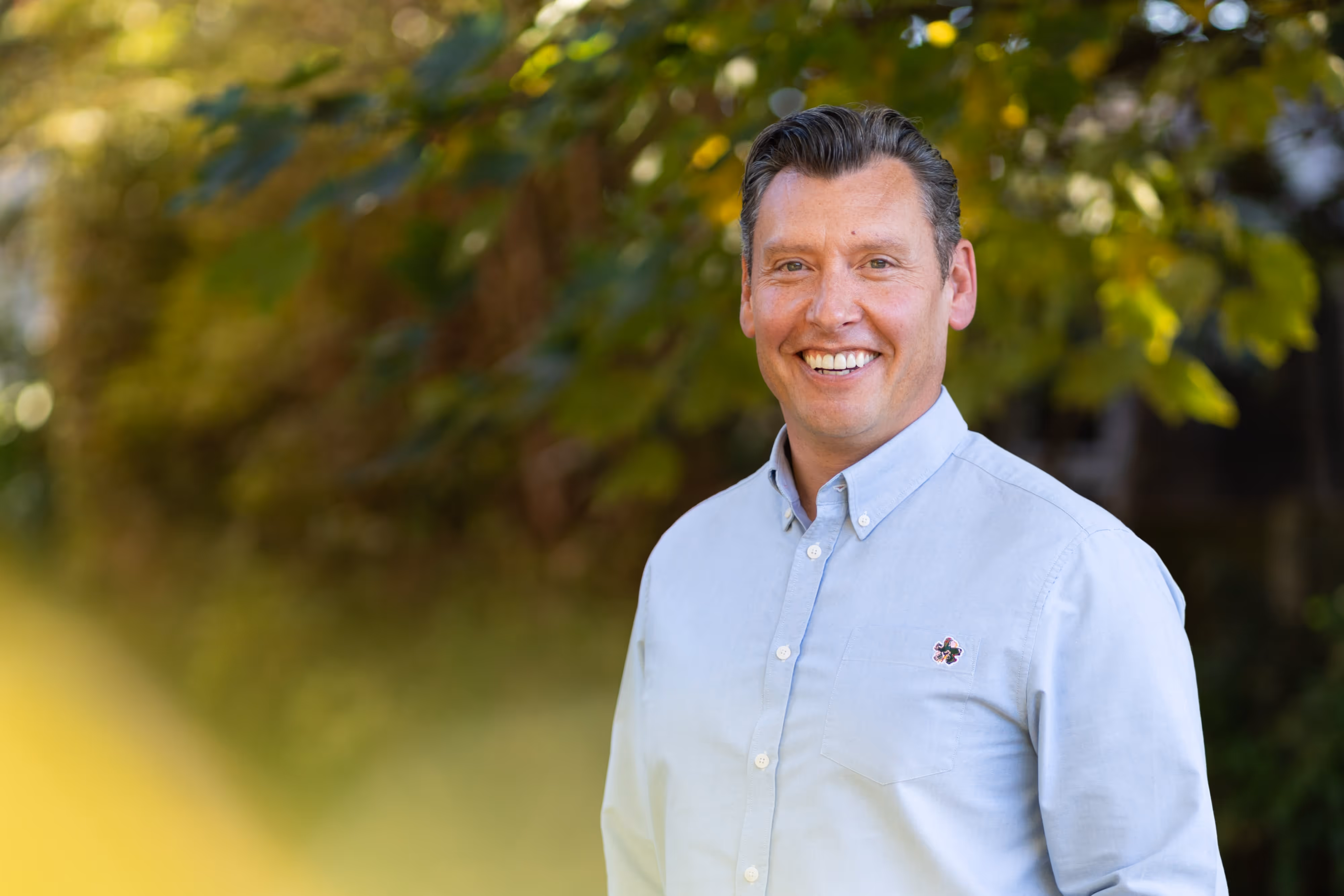 Smiling man in a light blue button-up shirt stands outdoors with green foliage in the background.