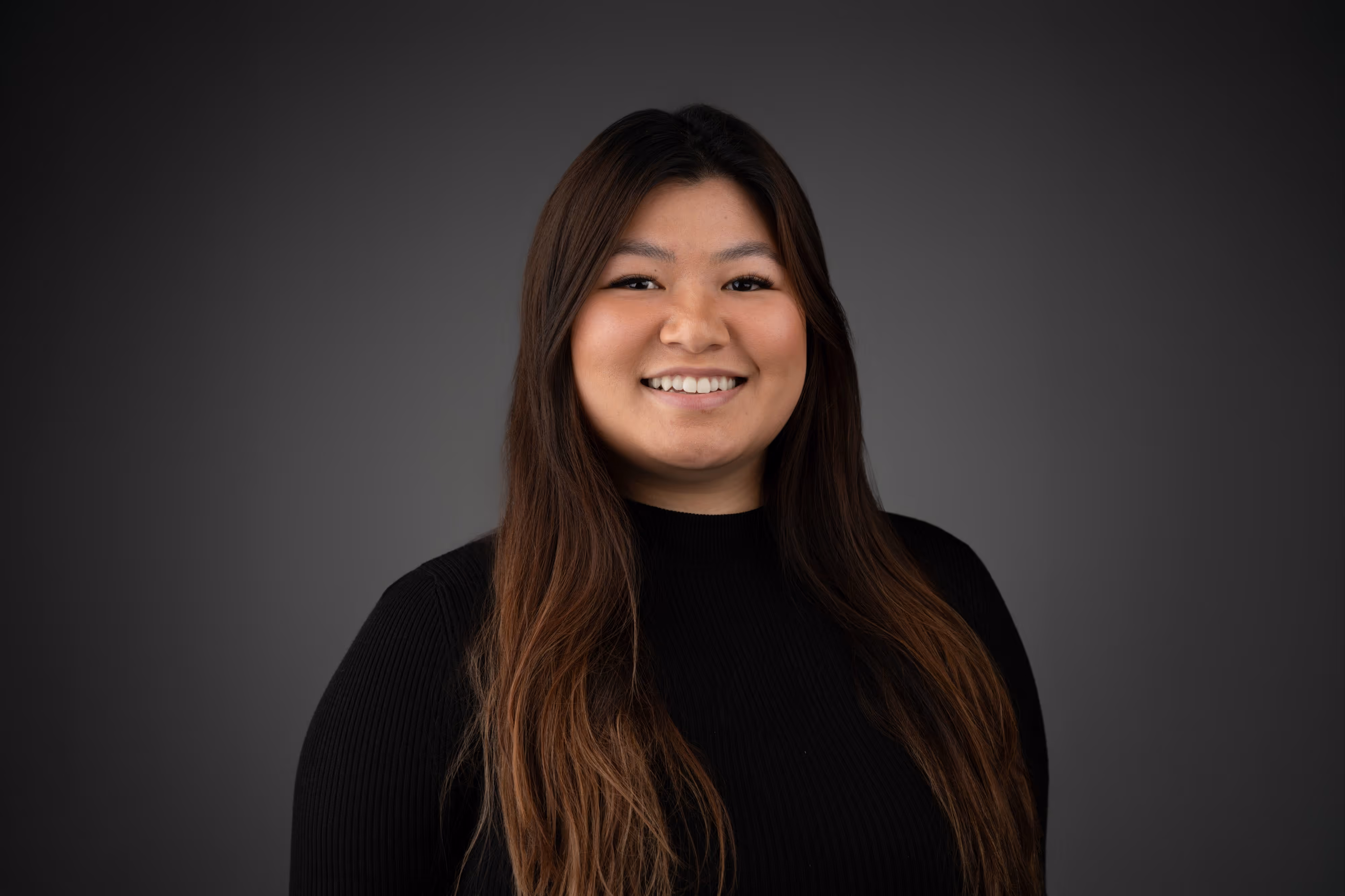 Woman with long brown hair wearing a black jumper, smiling in front of a dark grey background.