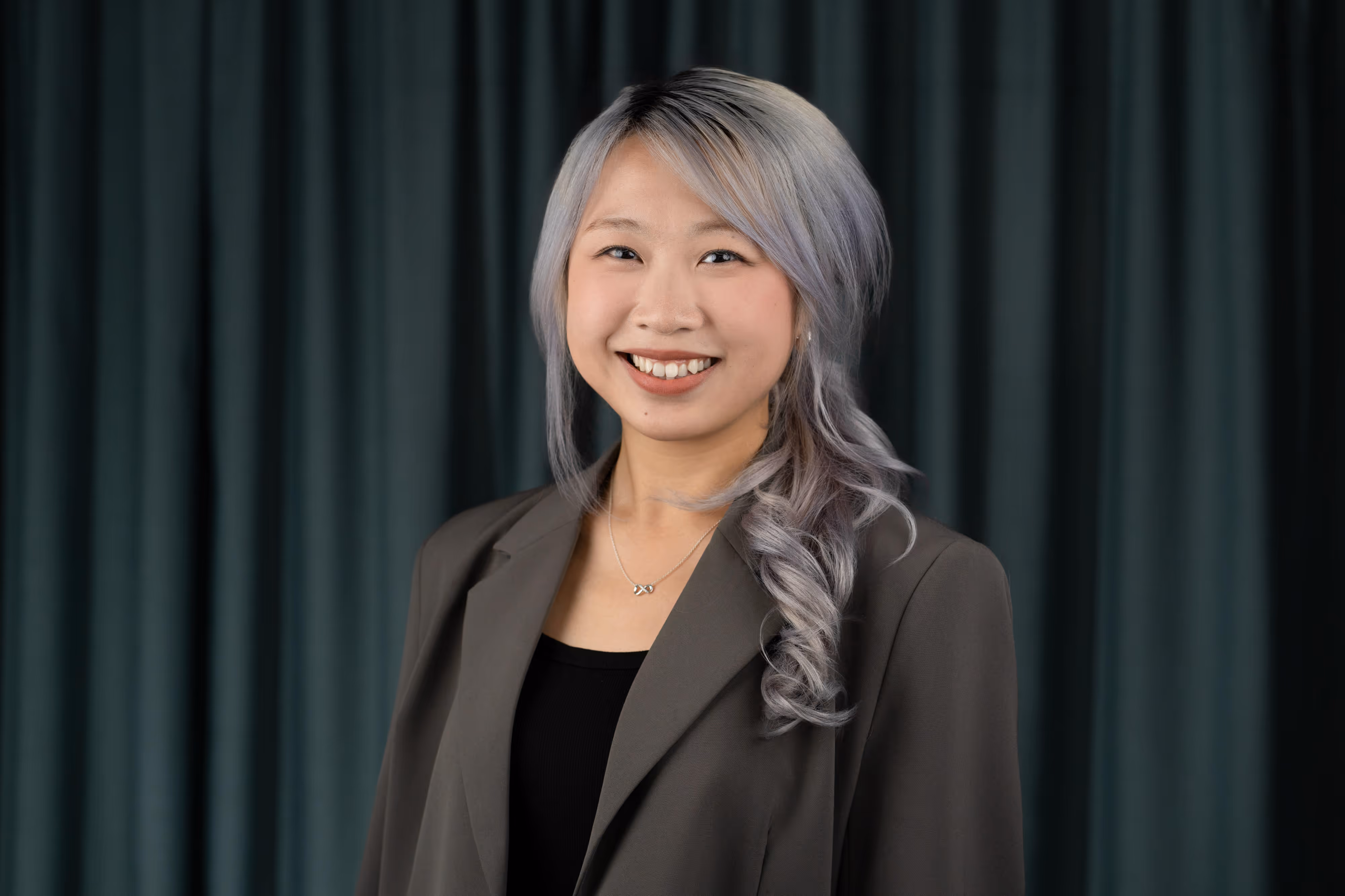 Smiling woman with long, wavy silver hair in a plait, wearing a dark blazer, standing in front of dark green curtains.