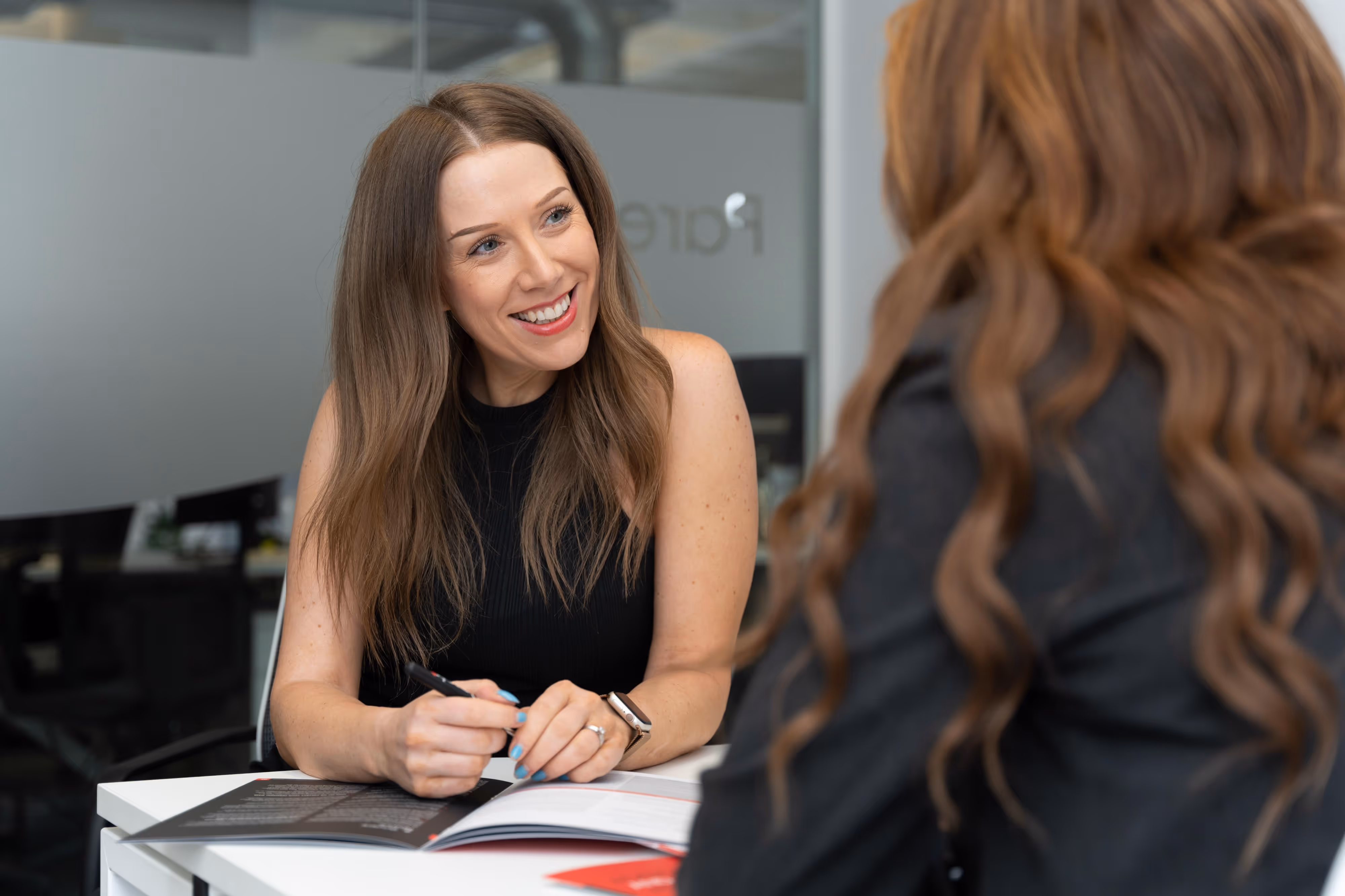 Two finance professionals sit at a table having a conversation, with papers and a pen in front of them in an office setting.