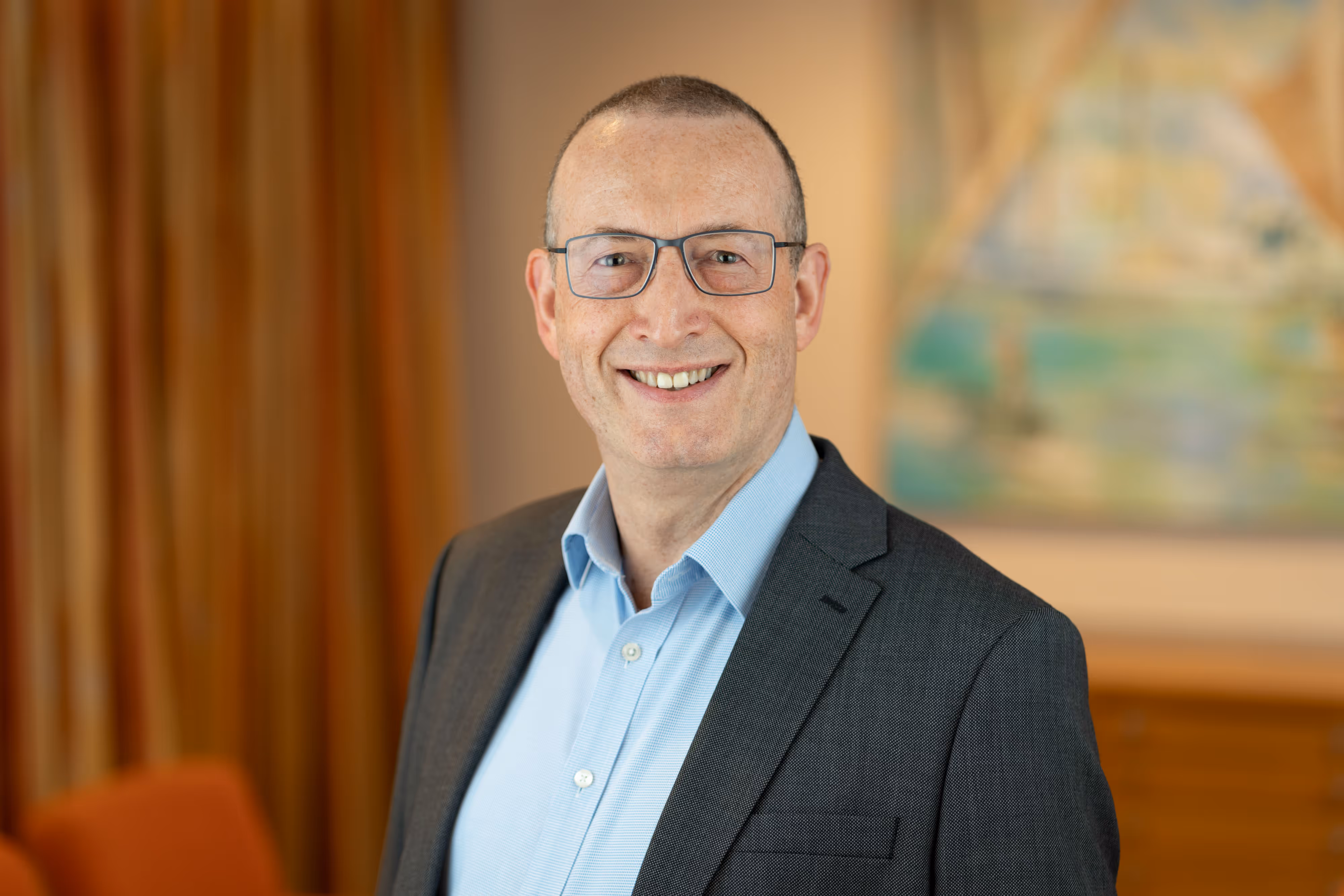 Smiling property professional in a suit and glasses standing indoors with a blurred background for a company headshot.