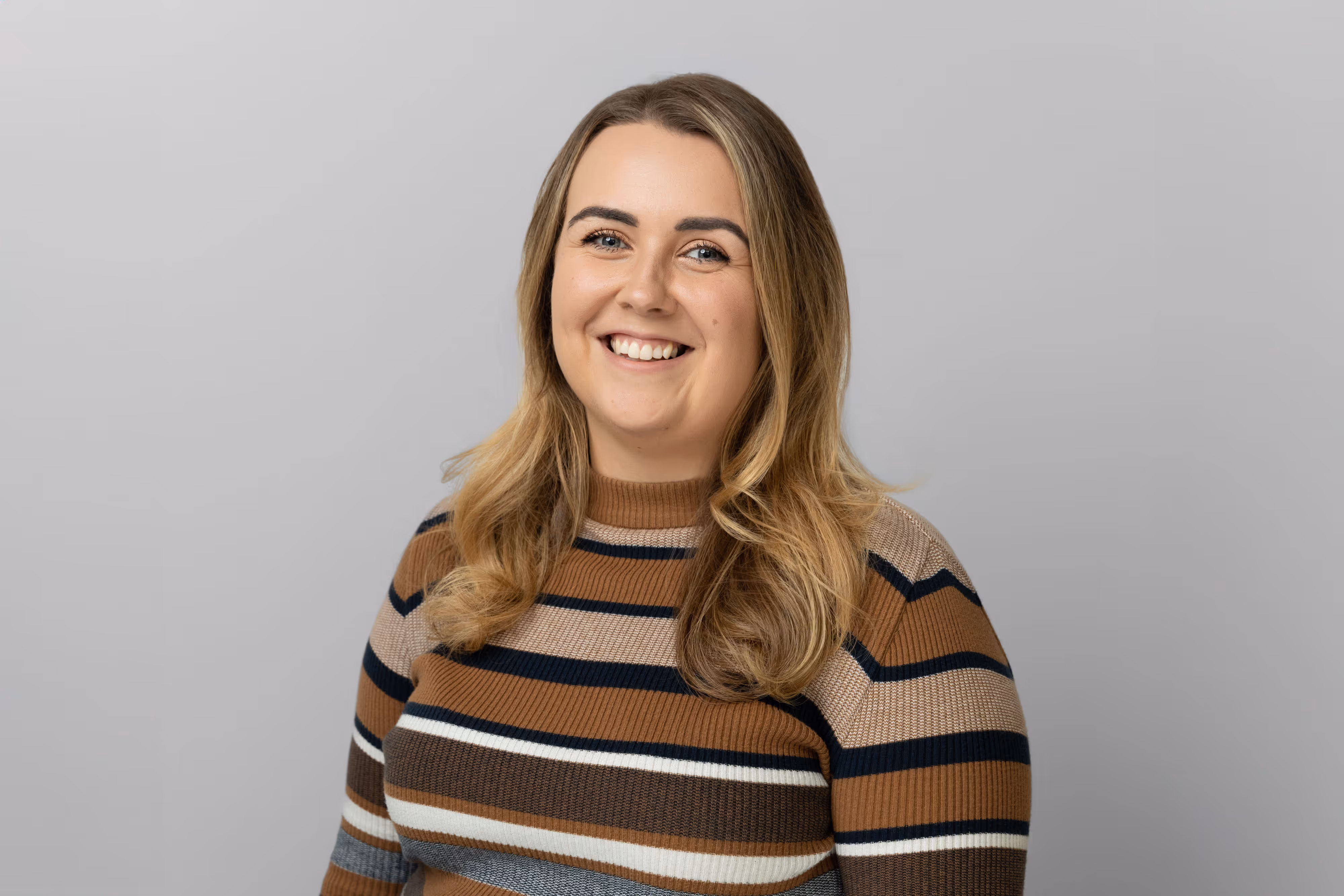 A woman with long, wavy blonde hair smiles, wearing a brown striped jumper against a plain light grey background.