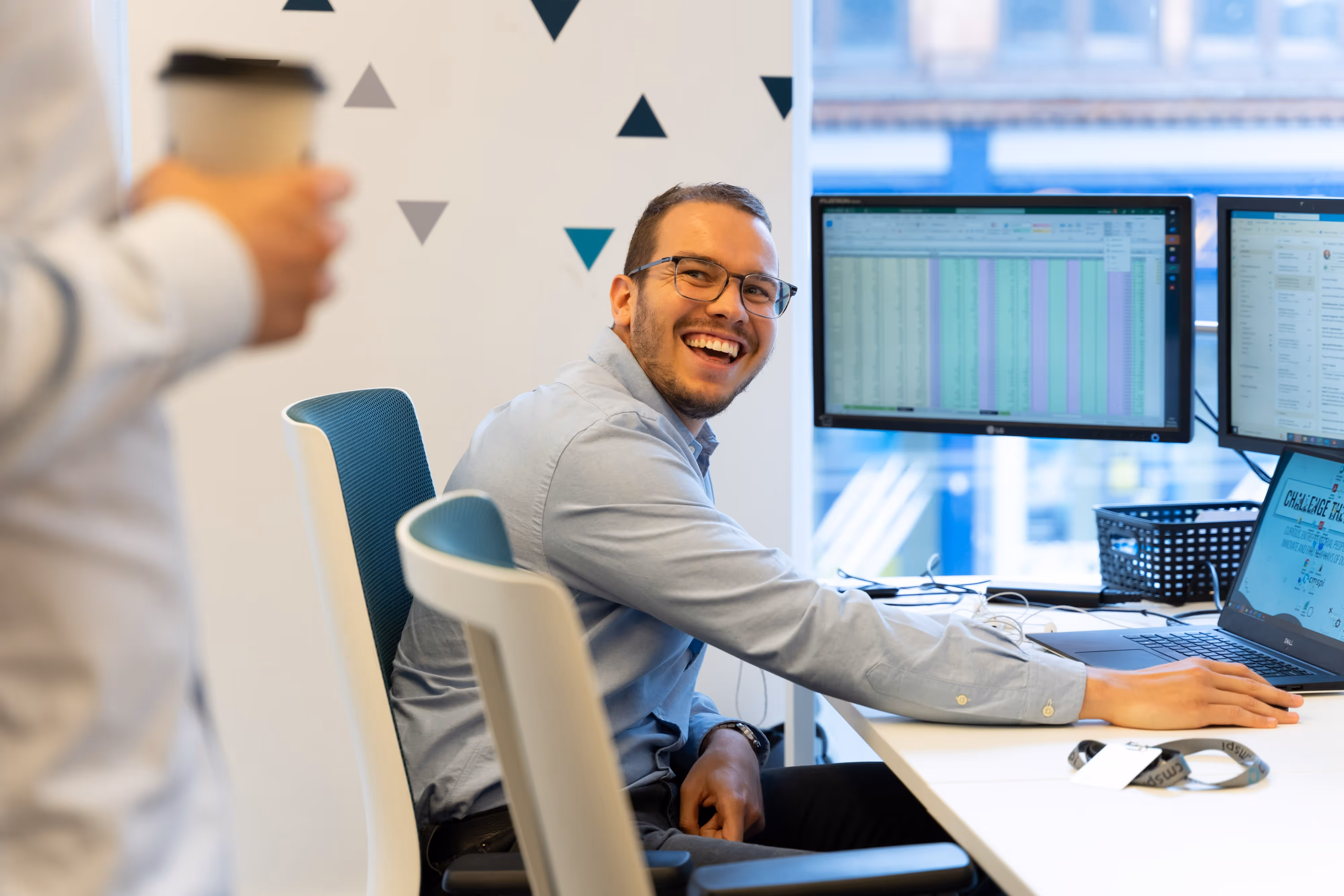 Smiling man at desk with computer monitors, looking up at a colleague holding a coffee cup in an office setting.