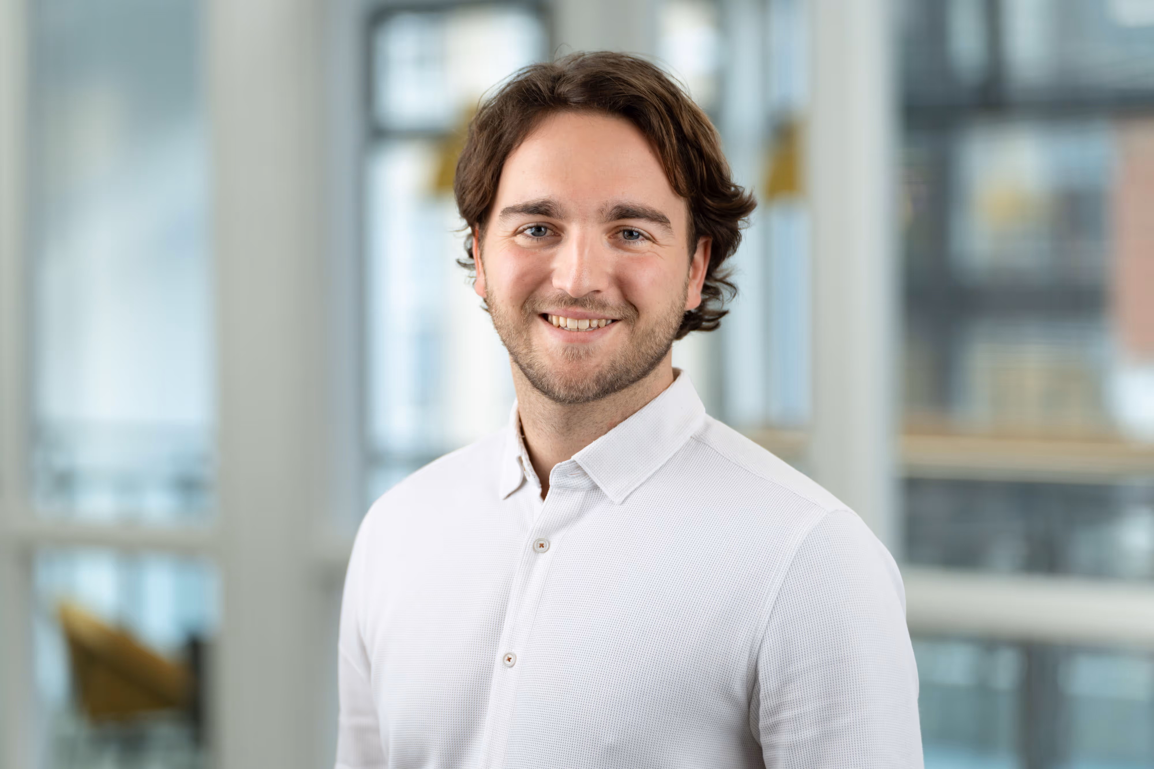 A young man with wavy brown hair smiles, wearing a white shirt, standing in a bright office setting.