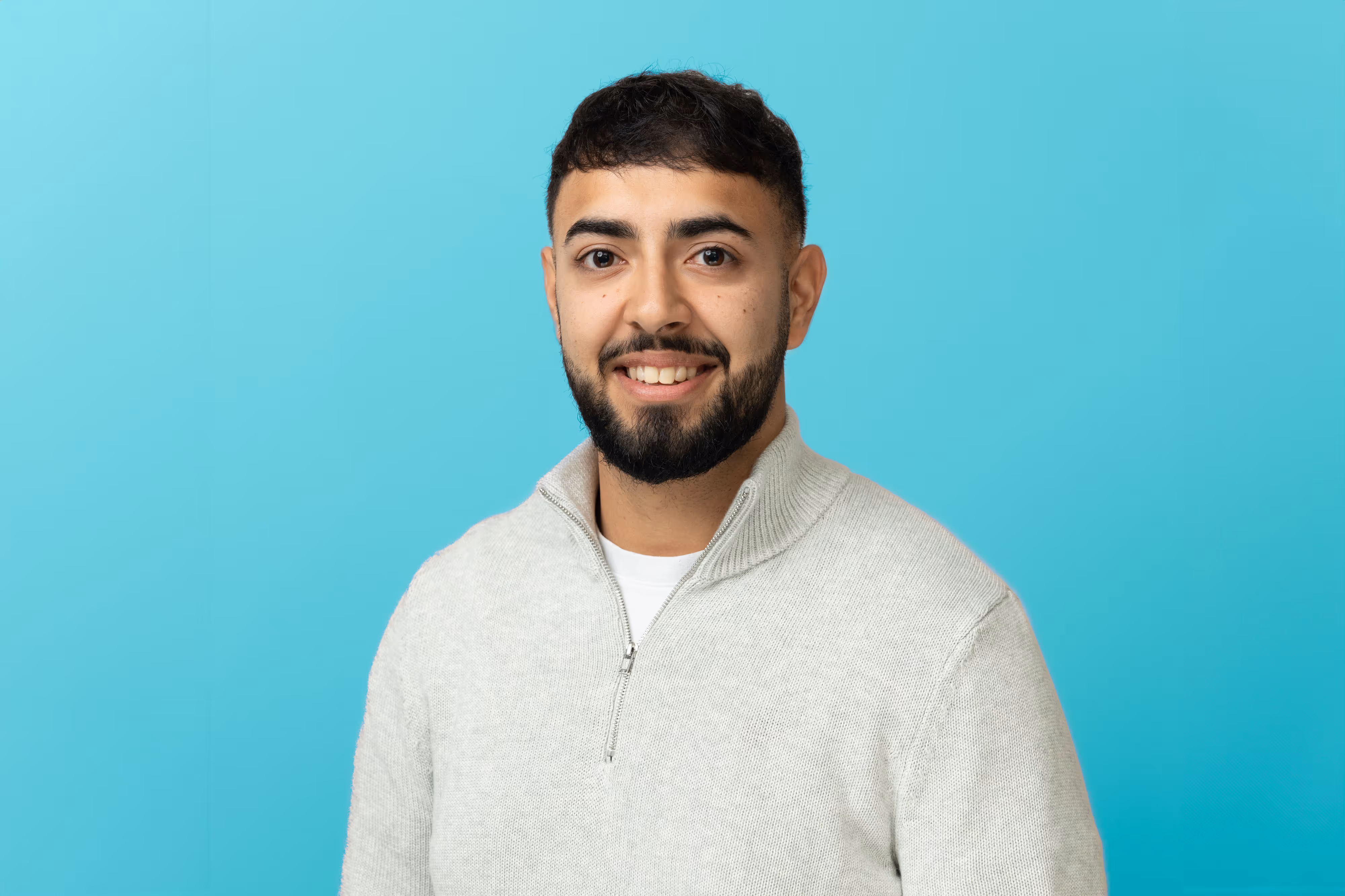 A smiling man with short dark hair and a beard, wearing a light grey jumper, stands against a blue background.