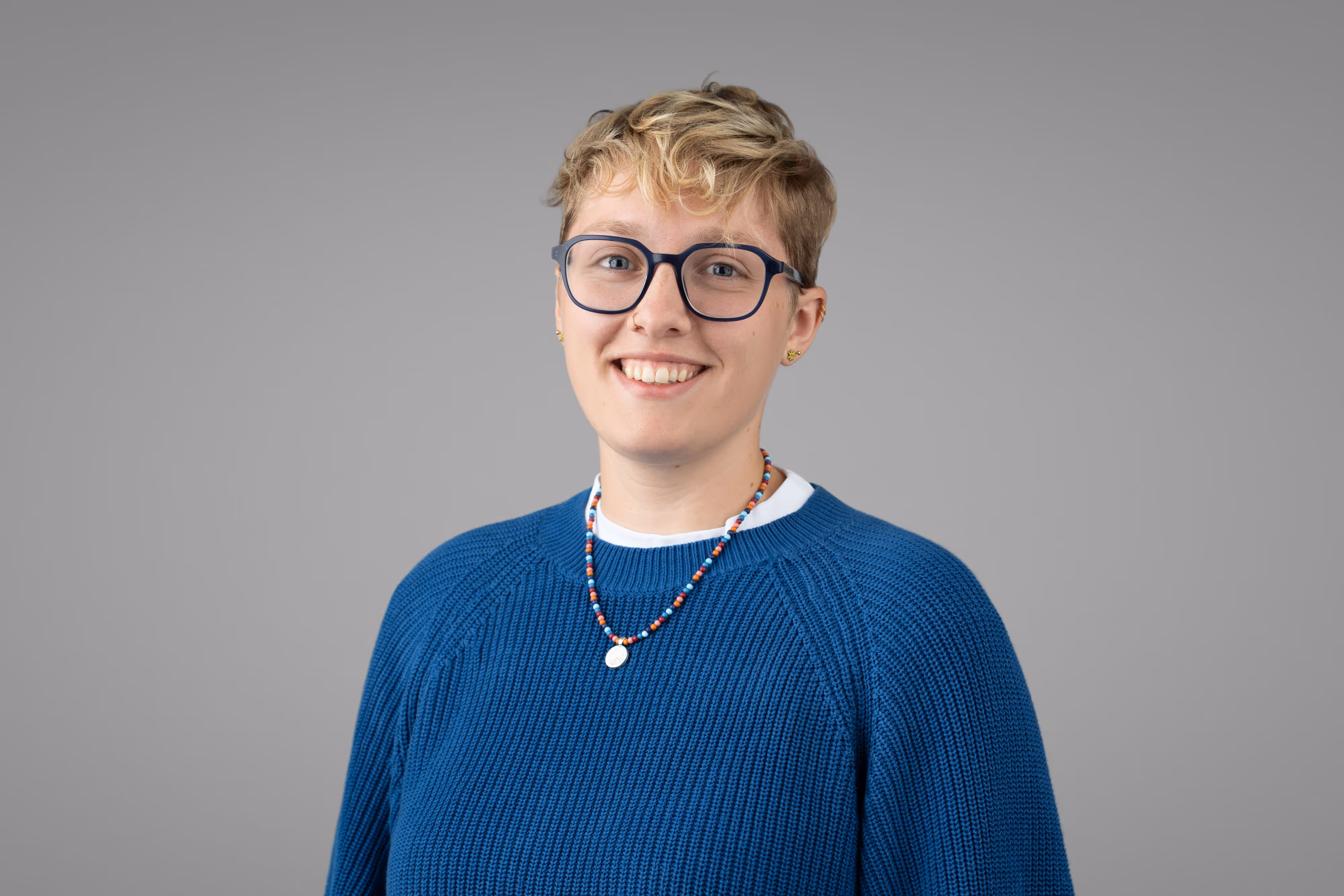 A person with short blond hair, glasses, and a blue jumper smiles at the camera against a plain grey background.