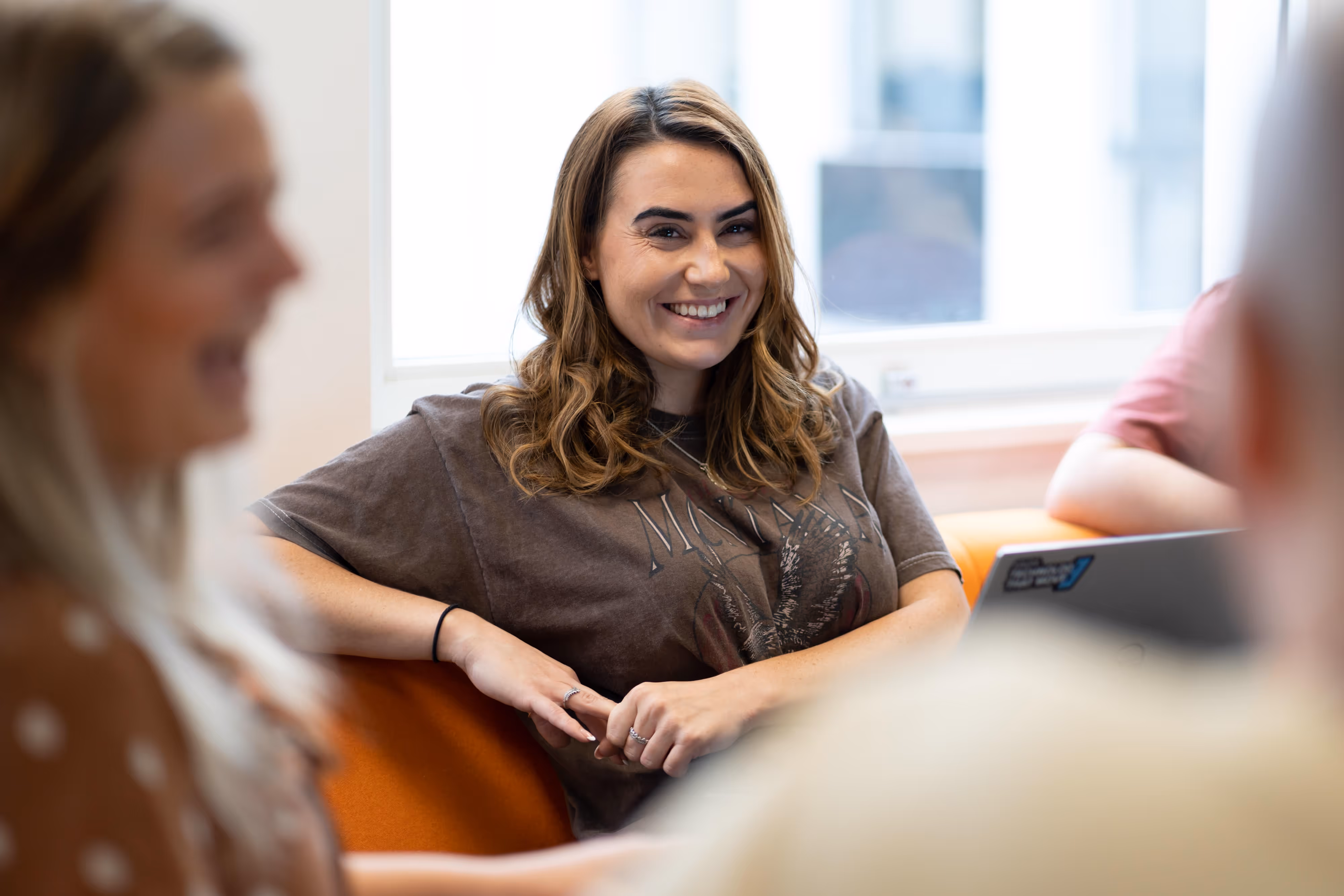 A woman with brown hair smiles candidly whilst sitting on an orange sofa, talking with others in a bright room.