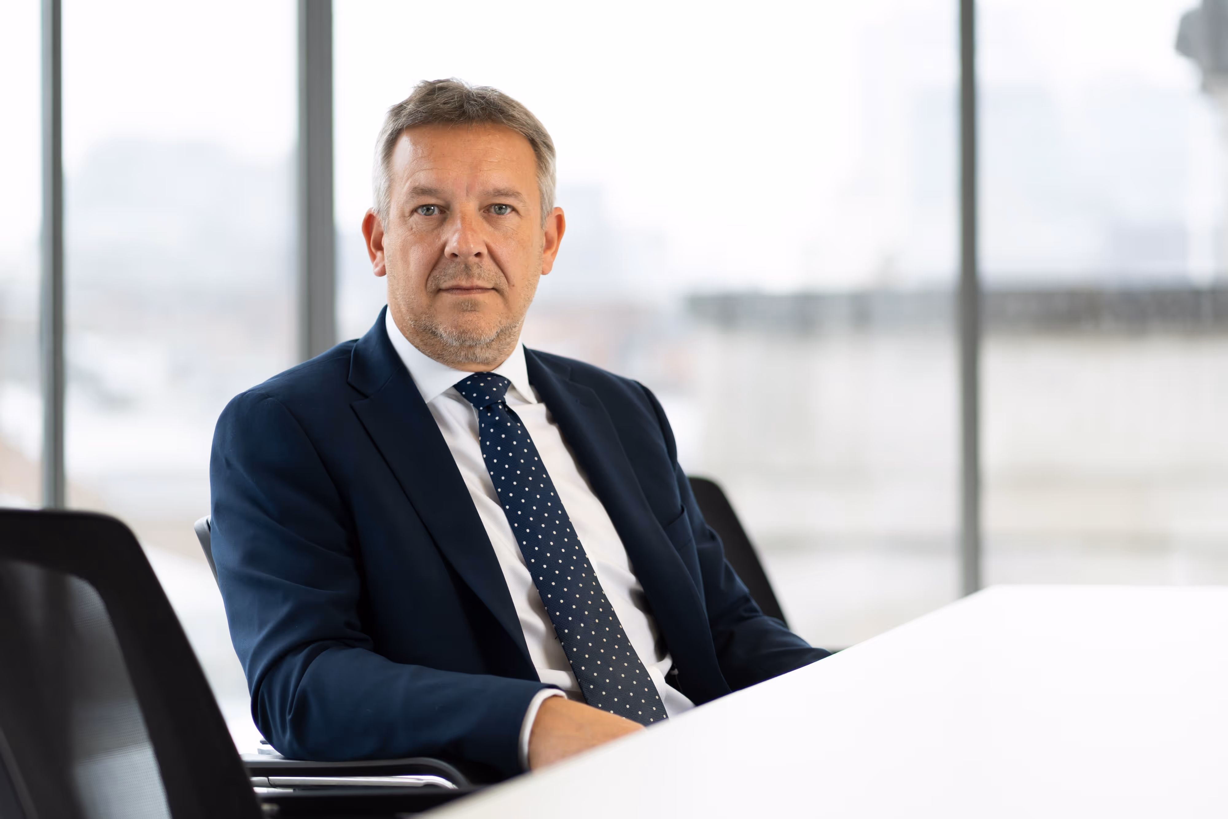 A man in a navy suit and polka dot tie sits at a white desk in a modern office with large windows.