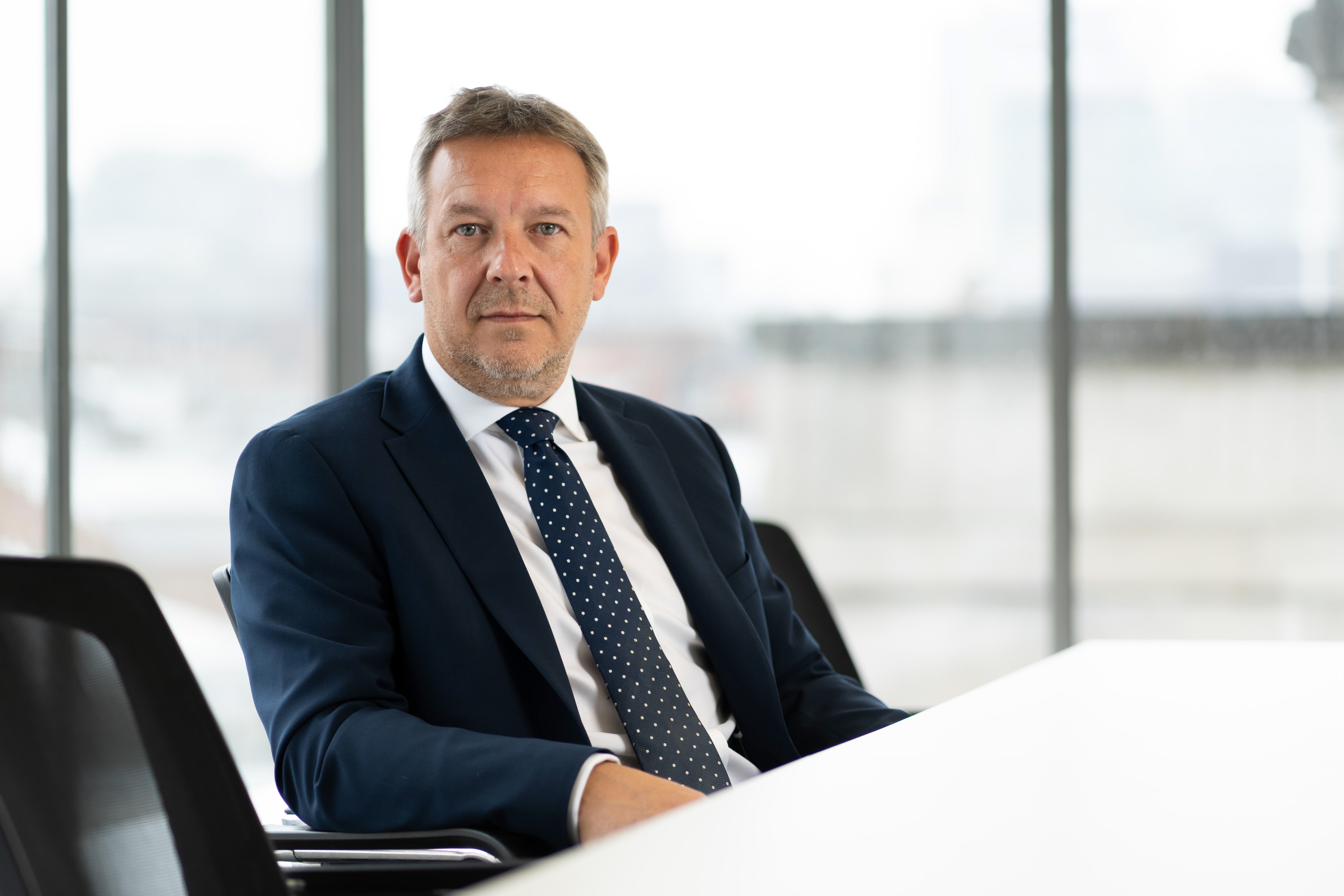 A man in a navy suit and polka dot tie sits at a white desk in a modern office with large windows.